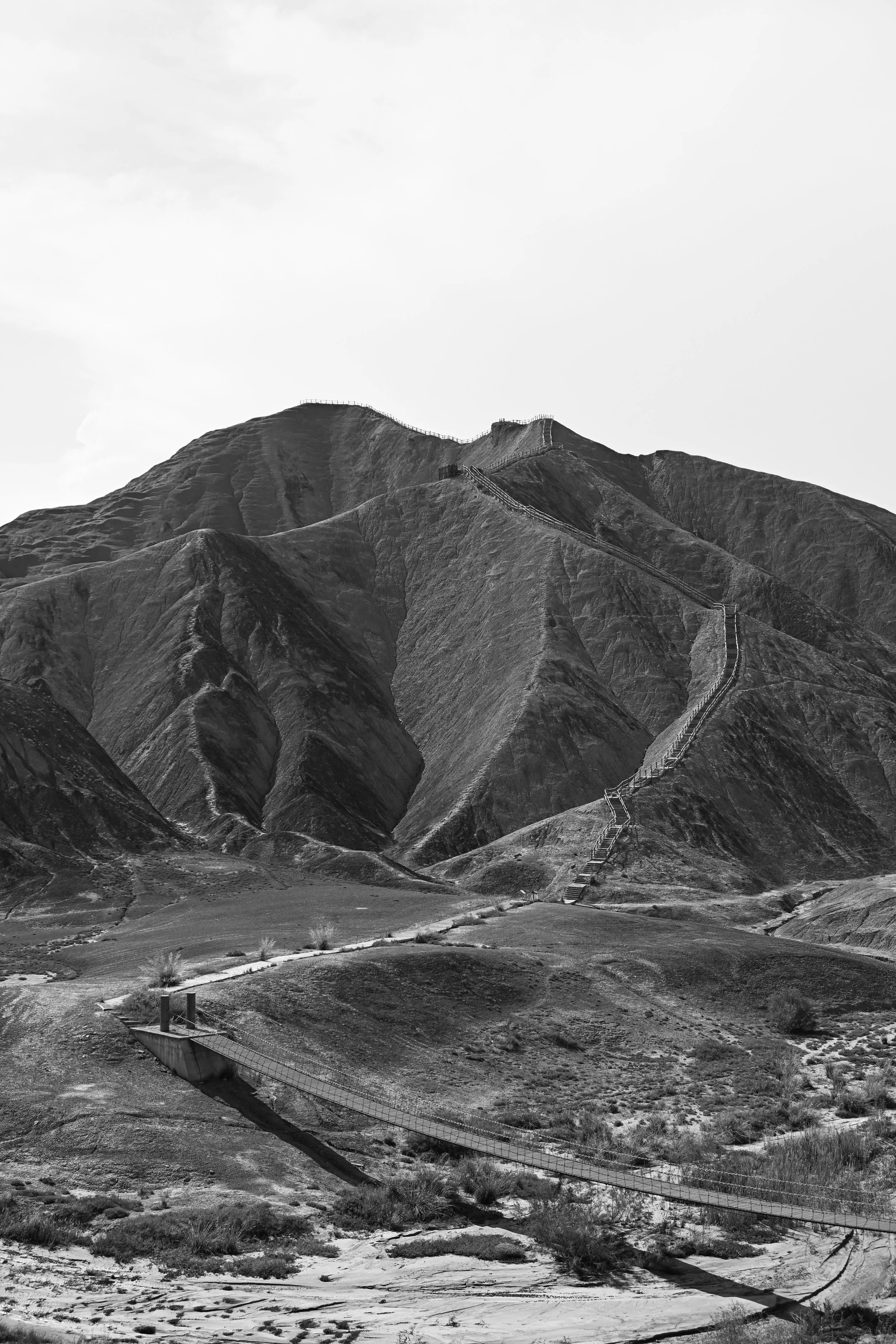 Black and white photo of a mountain landscape with a long staircase climbing the mountain, and a bridge at the bottom.