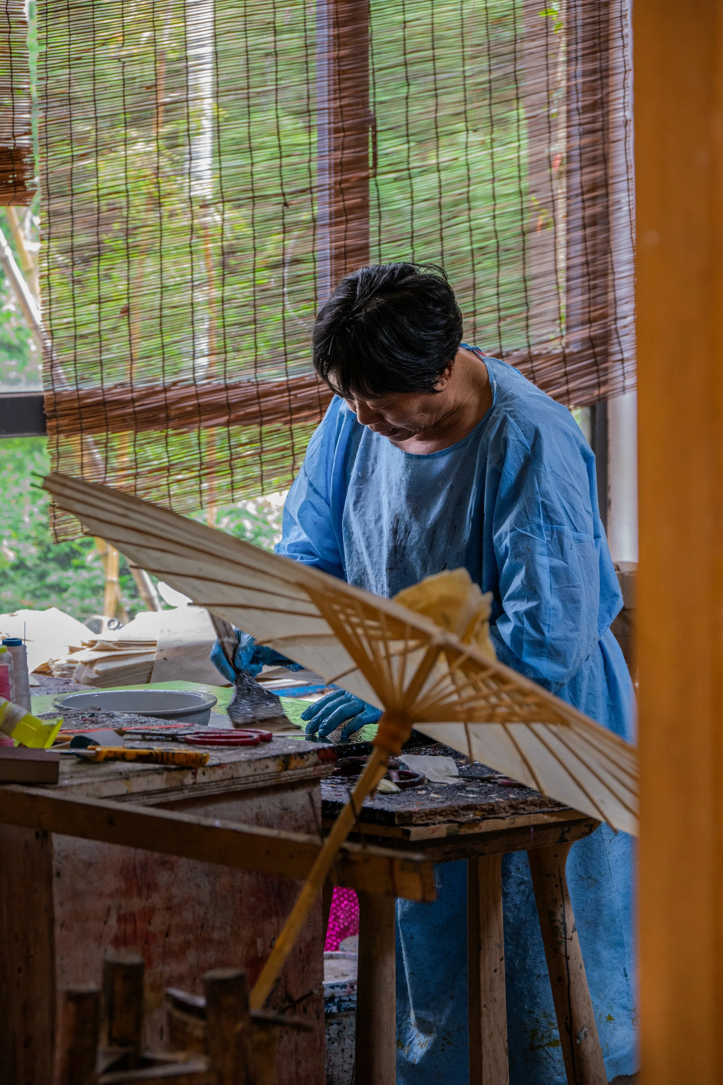 A person wearing a blue smock and gloves working on a craft project at a cluttered wooden worktable, with a traditional paper umbrella and bamboo blinds in the background.