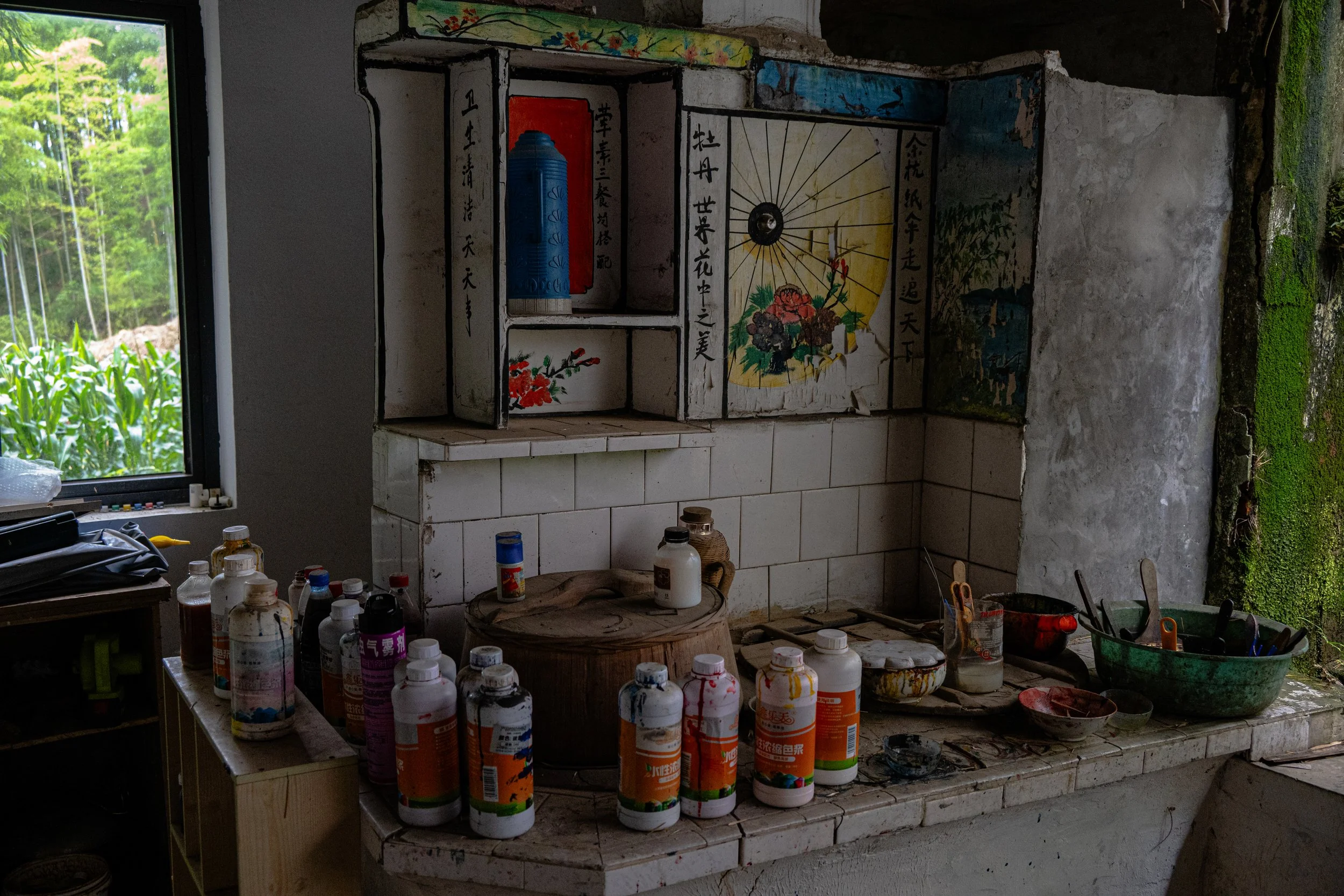 An indoor area with a window looking out to a green field of corn. The space has a tiled surface with bottles, containers, and kitchen utensils scattered on it. There is a decorative, painted wooden cabinet or shelf mounted above the tiles with Chine