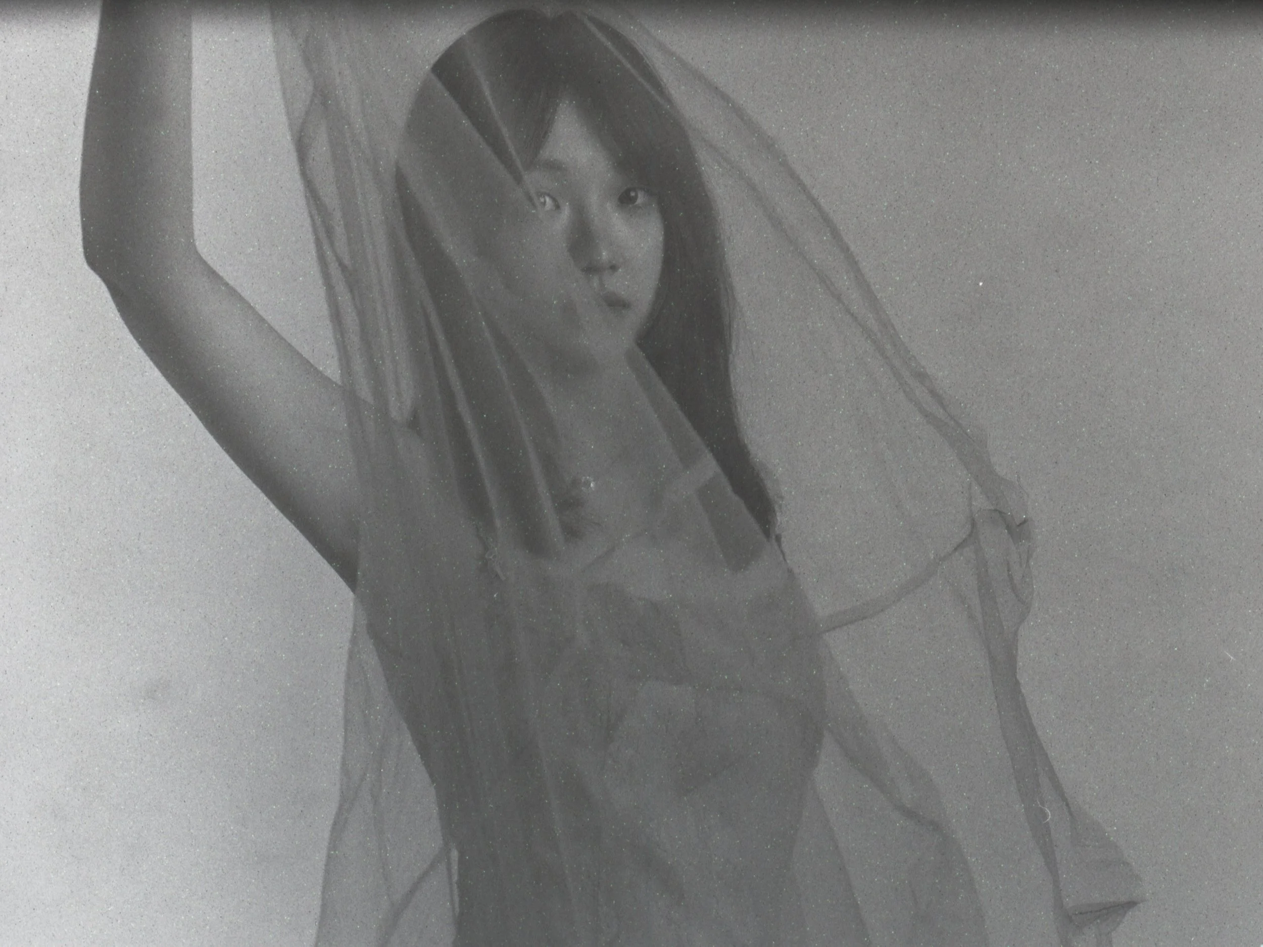 Woman with long hair holding transparent fabric above her head, looking at camera in gray-scale photo.