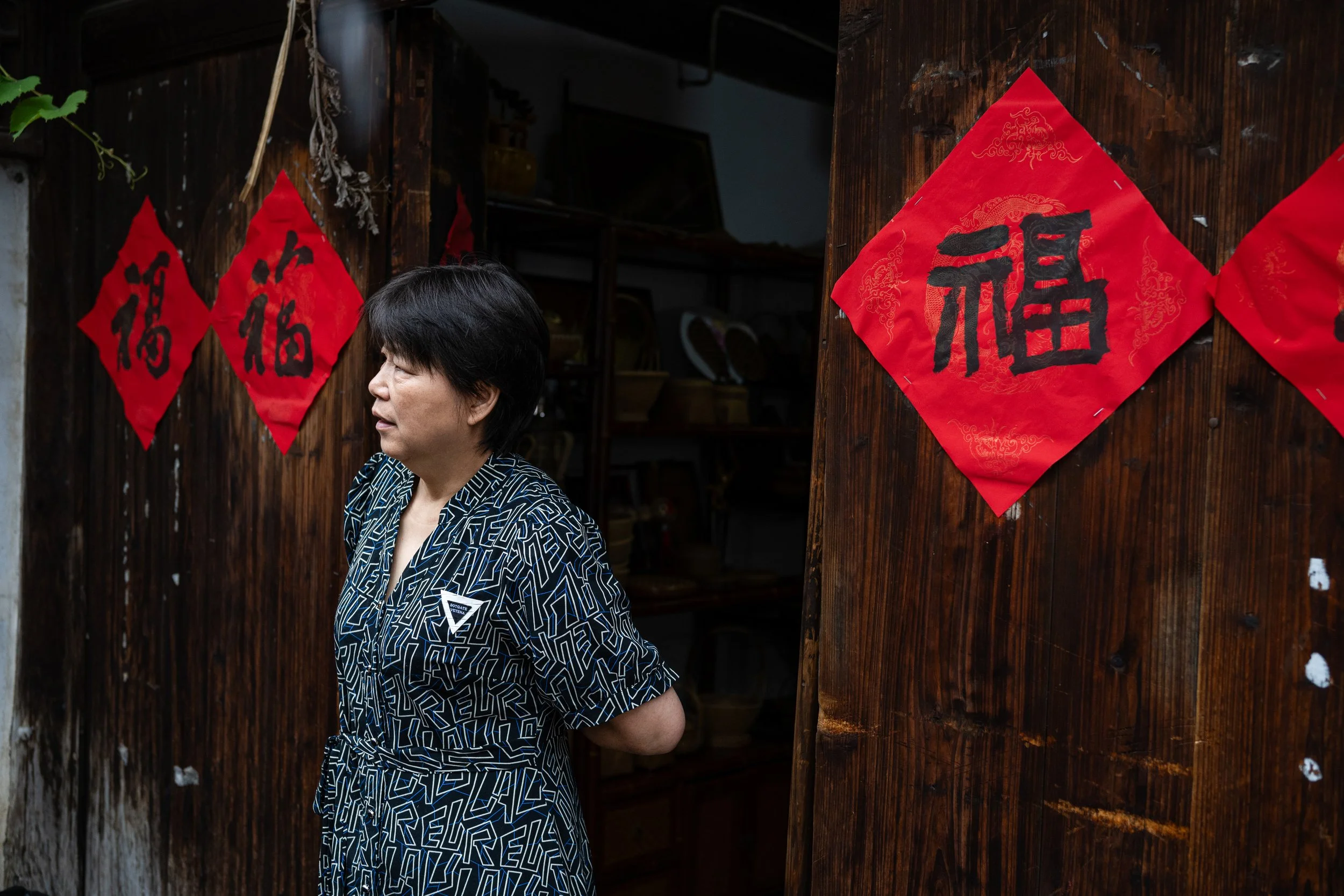 A woman standing indoors in front of a wooden wall decorated with red Chinese New Year couplets with black calligraphy.