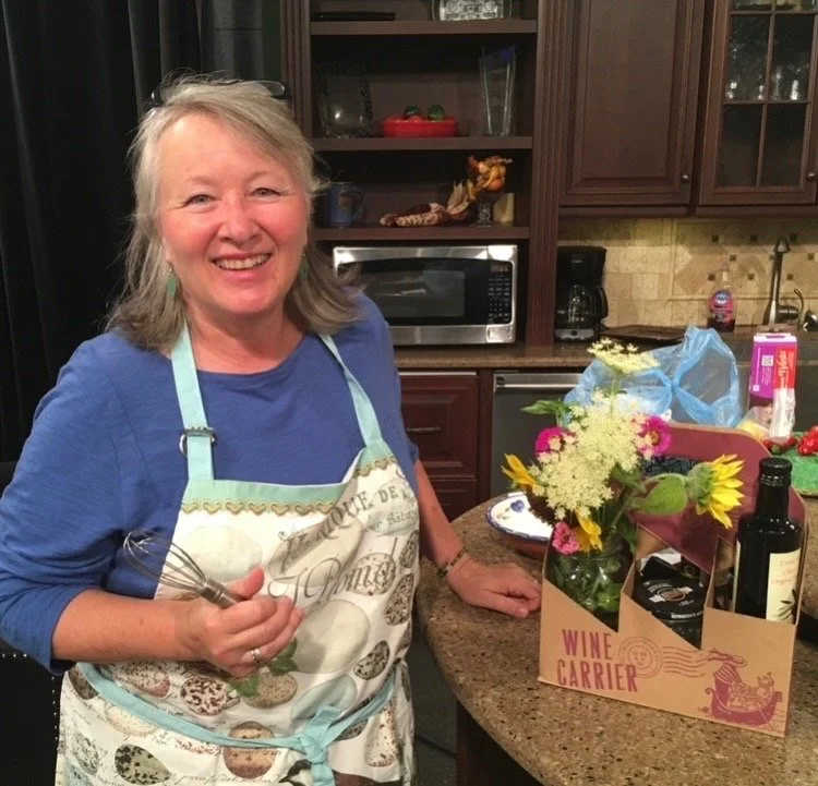 A woman with gray hair smiling in a kitchen, holding a whisk and wearing a floral apron over a blue shirt, next to a flower arrangement and bottles on a kitchen counter.