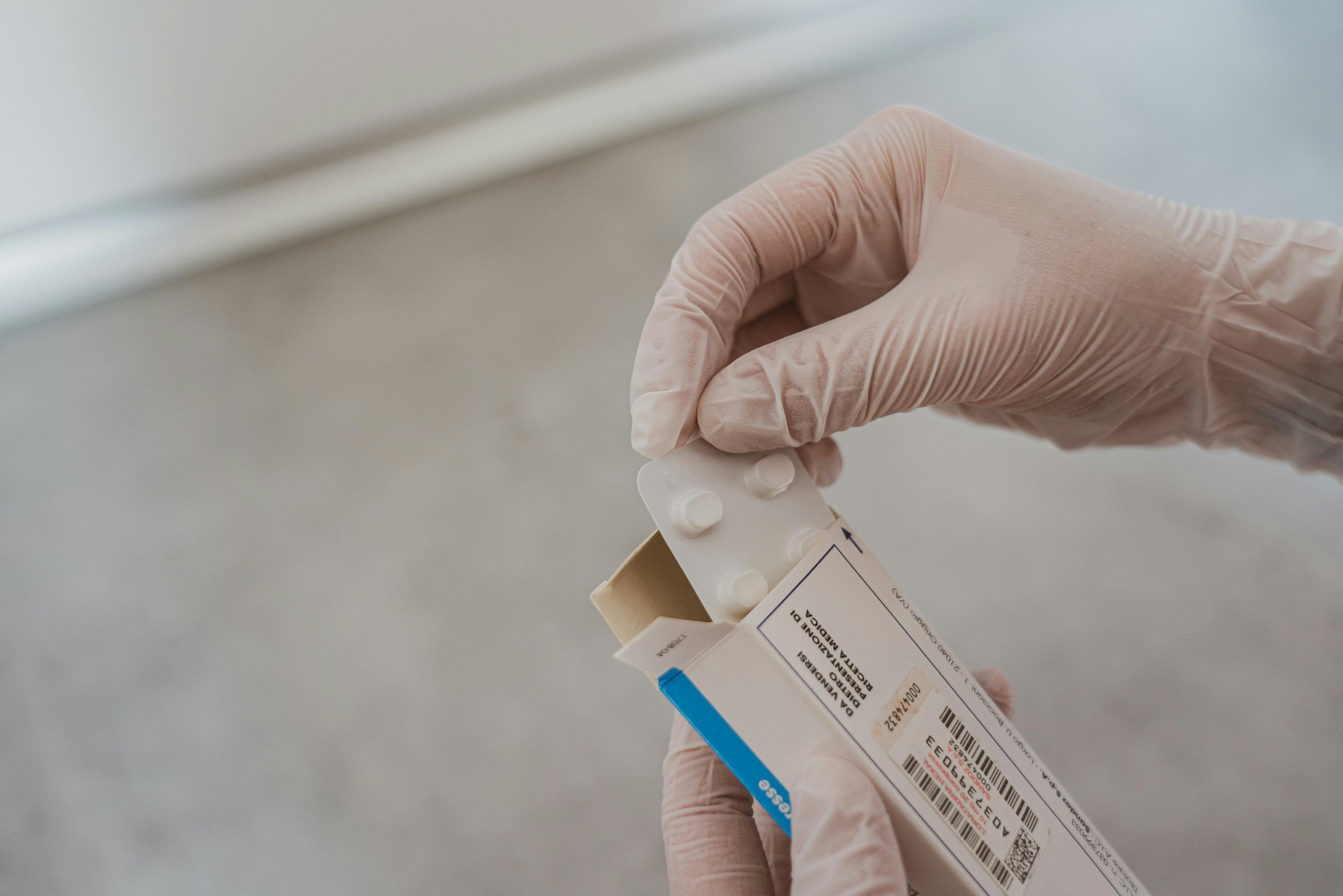 A gloved hand holding a box of medication with a blister pack of tablets being removed.