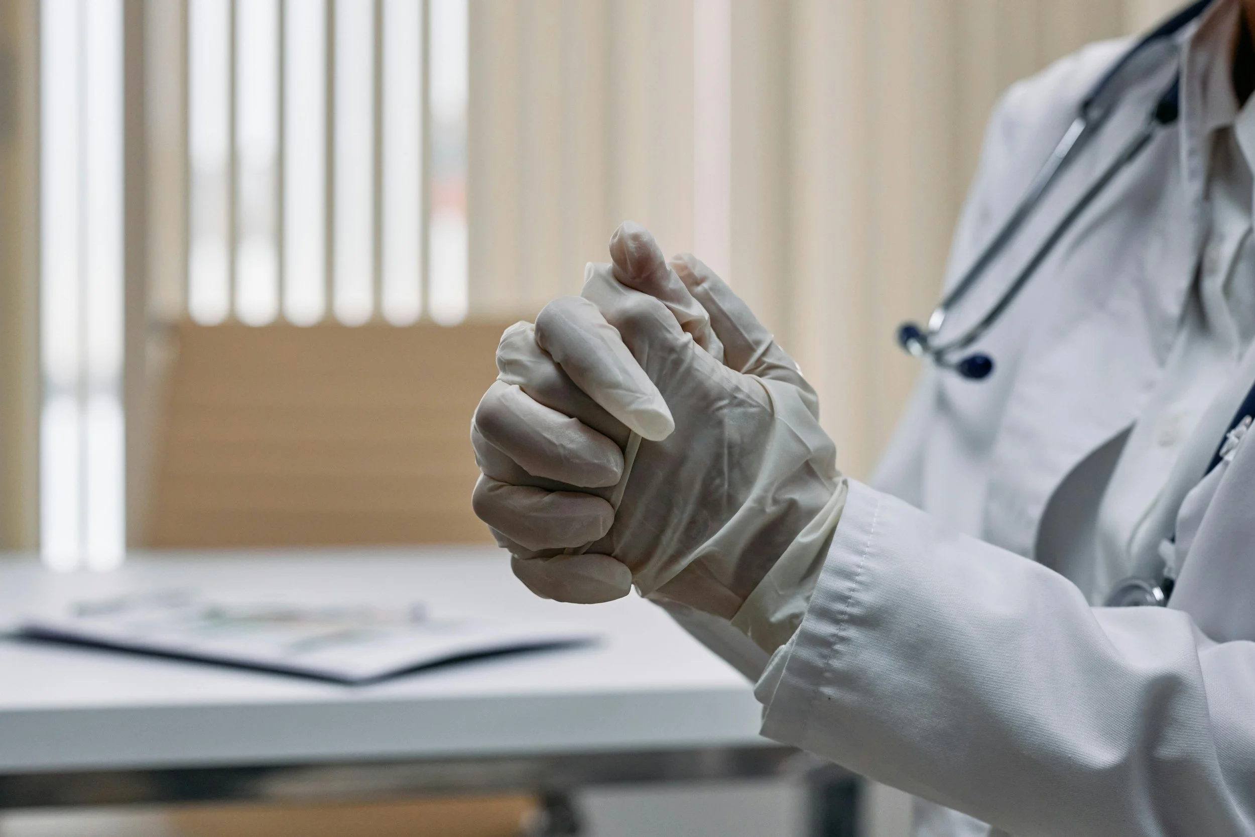 Close-up of a doctor wearing white gloves and a white coat, with a stethoscope around their neck, sitting at a desk.