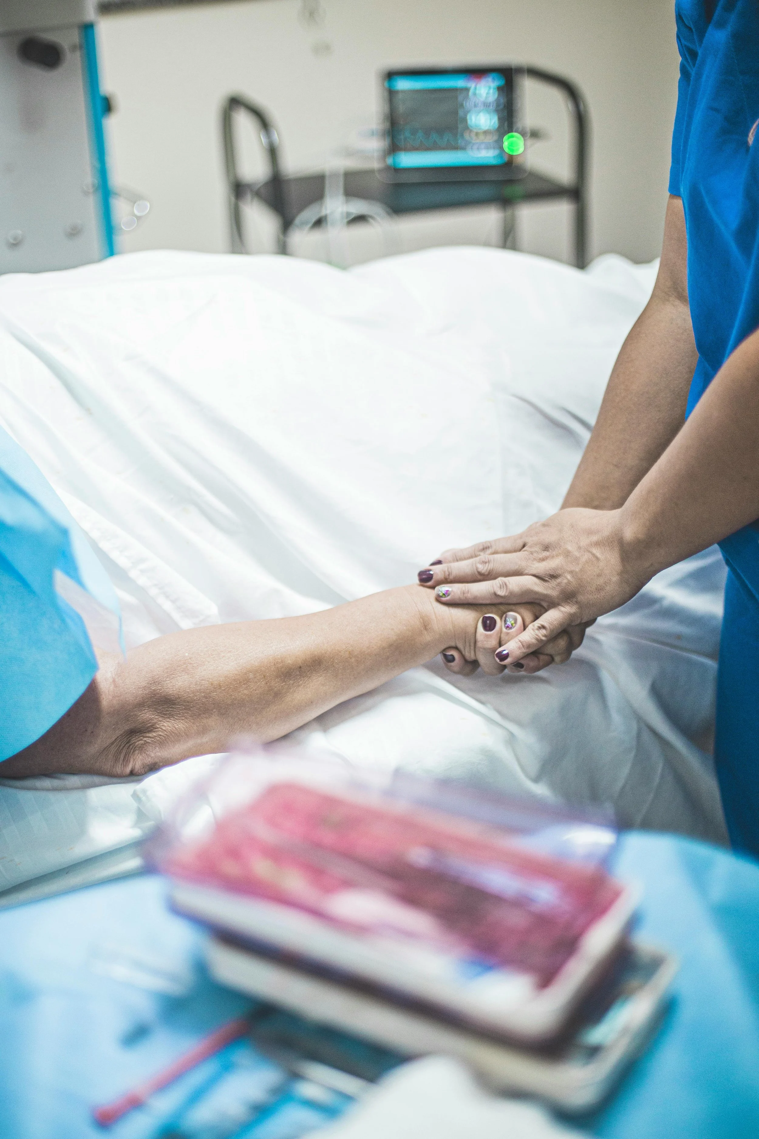 A healthcare professional holding a patient's hand in a hospital setting with medical monitors in the background.