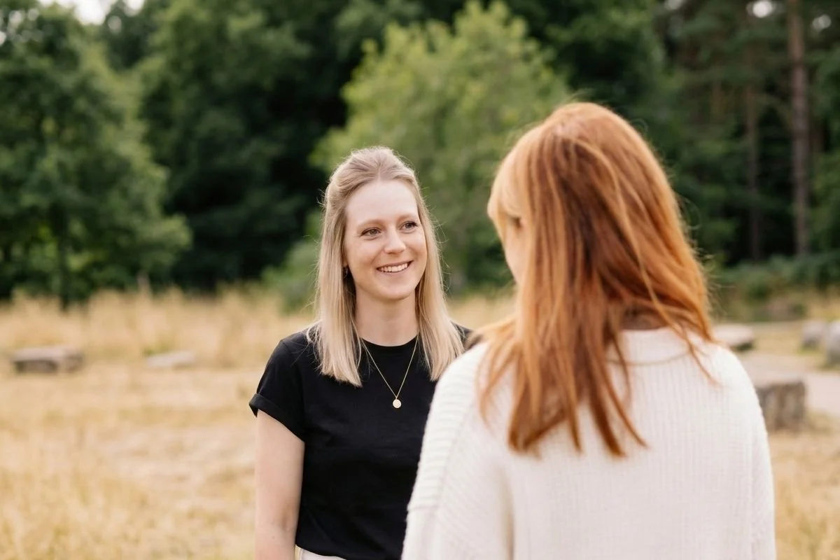 Zwei Frauen sprechen im Freien, eine lächelt und trägt schwarze Kleidung, die andere mit roten Haaren trägt eine hellfarbige Pullover. Im Hintergrund sind Bäume und Gras zu sehen.