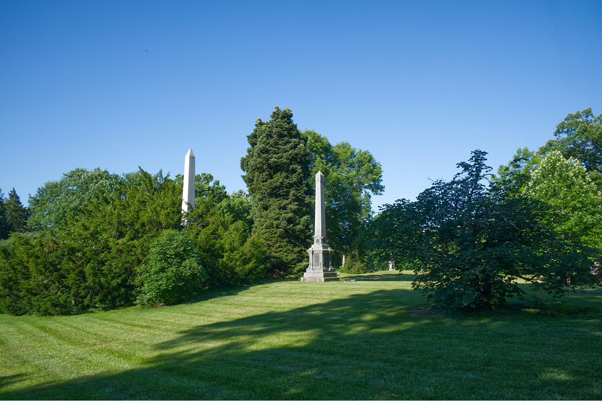 A park with green grass, tall trees, and monuments with obelisks under a clear blue sky.