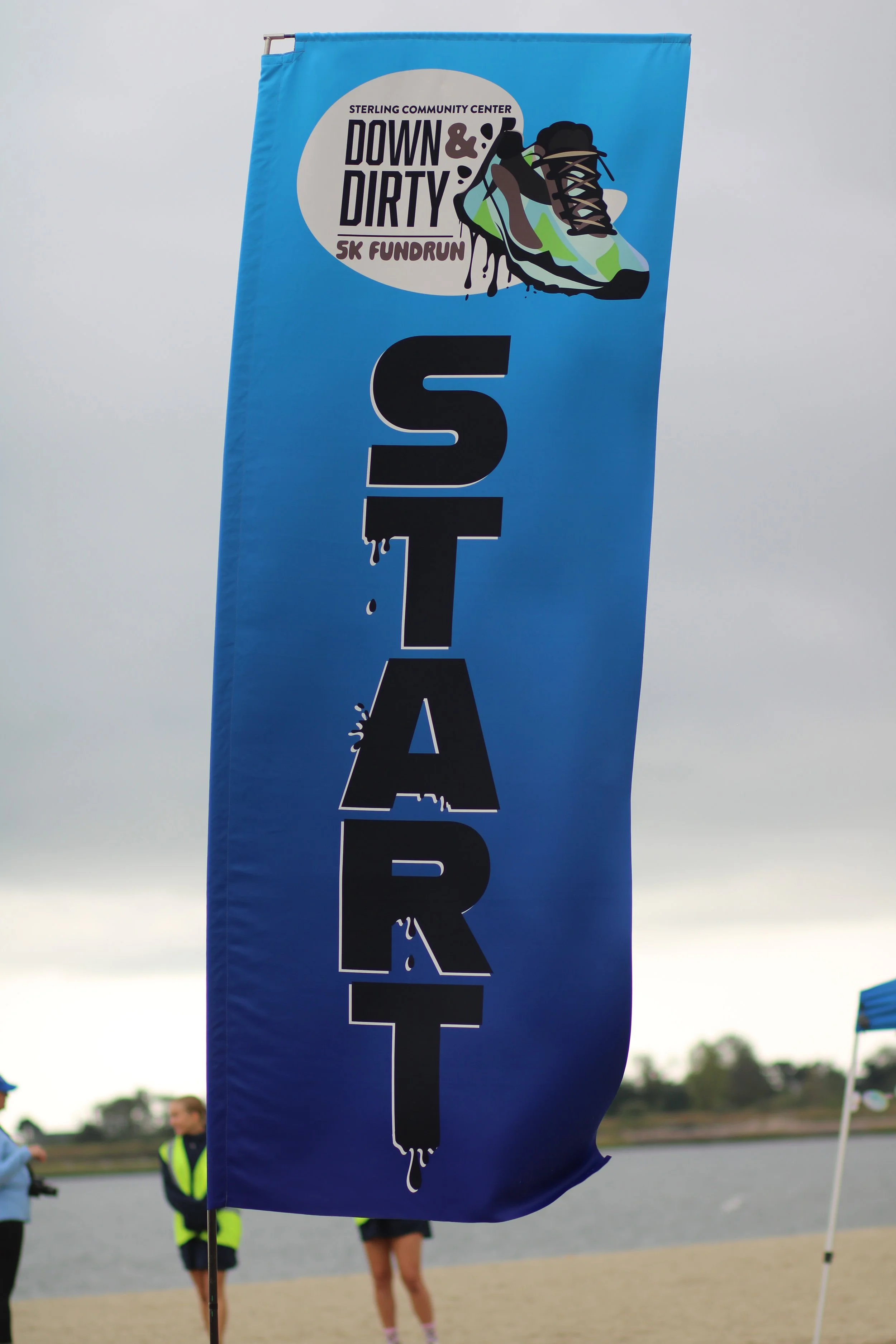 Blue rally flag promoting the Down & Dirty 5K Fun Run with a graphic of a running shoe and text, set against a lakeside background on an overcast day.