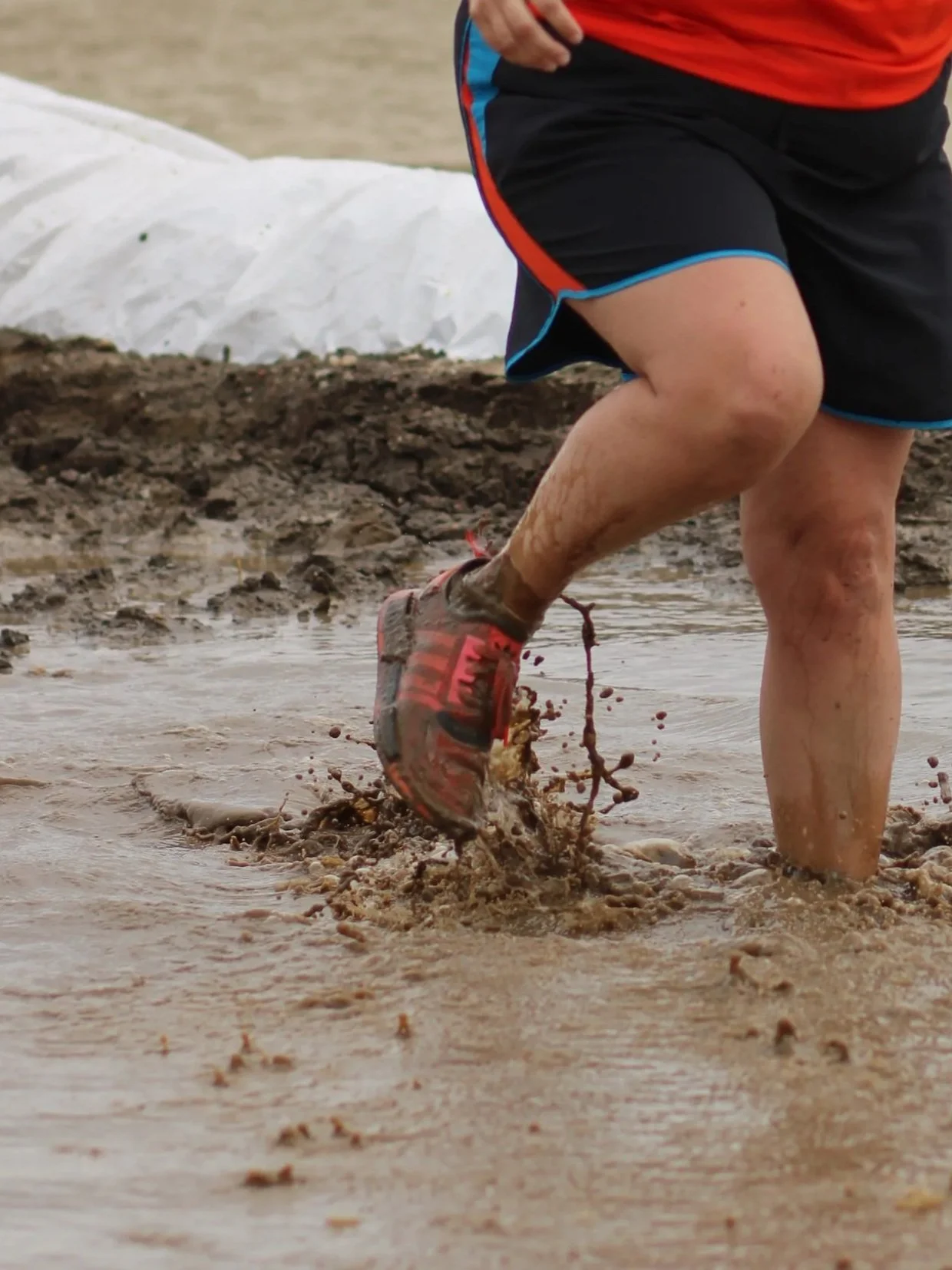 Person running through muddy water, wearing athletic shoes and shorts, with mud splashing around.