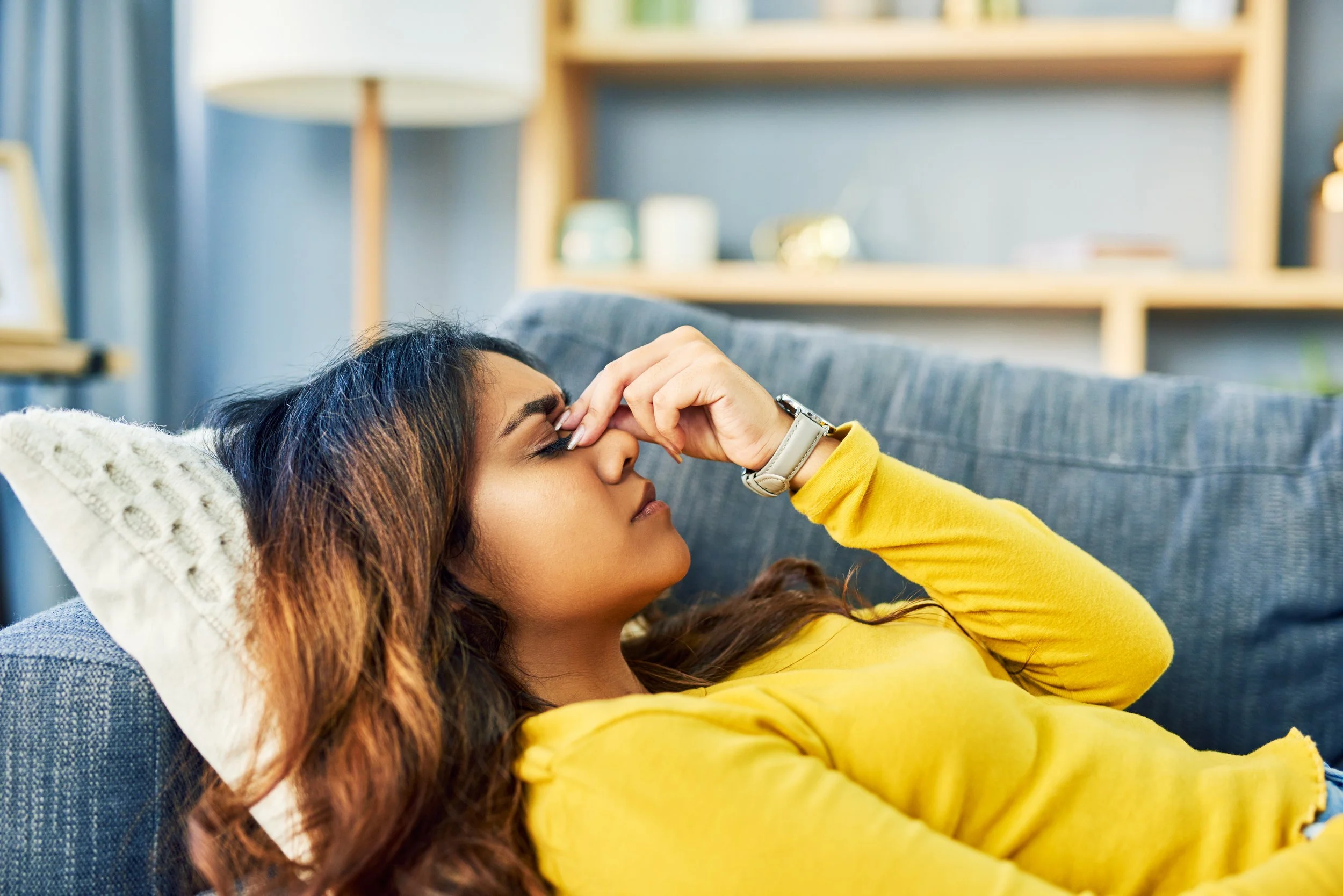 A woman with long, wavy hair lying on a blue couch, resting her head on a knitted pillow, wearing a yellow long-sleeve top, with her eyes closed and hand touching her forehead.