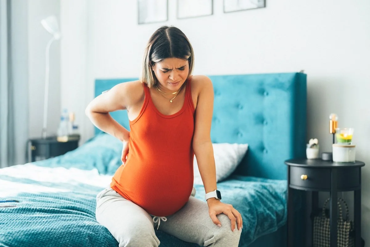 Pregnant woman sitting on bed, holding her lower back, appearing to experience discomfort or pain.