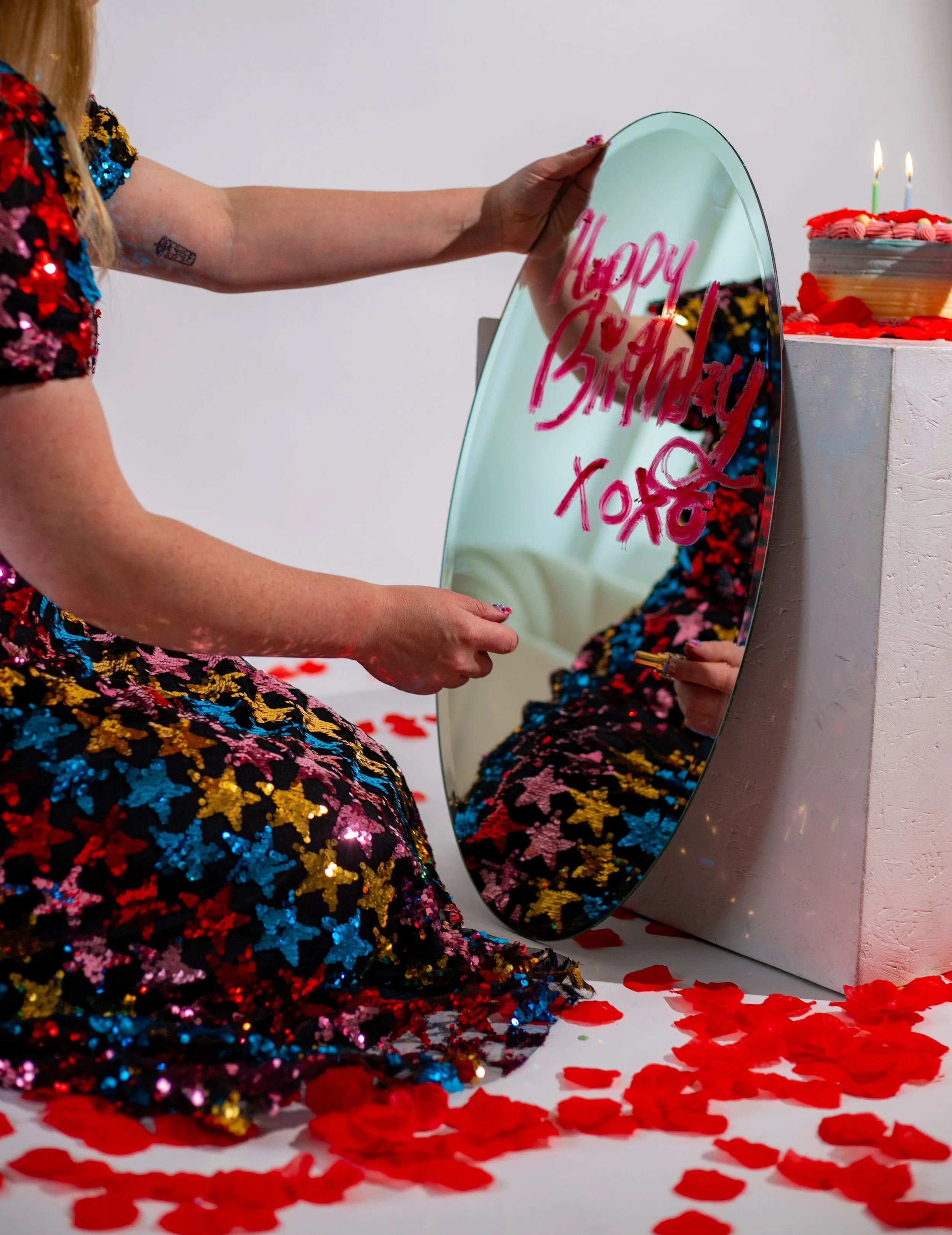 Person in a colorful star-patterned dress writing on a mirror with a pink marker, with a birthday cake and candles in the background.