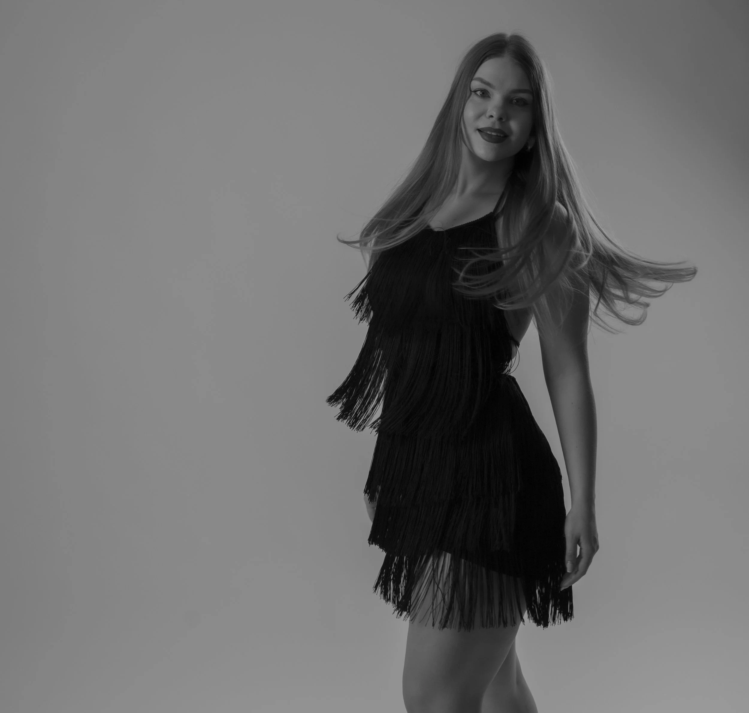 A woman with long hair wearing a black dress with fringe details, looking at the camera and smiling, with her hair flowing to the side against a plain background.