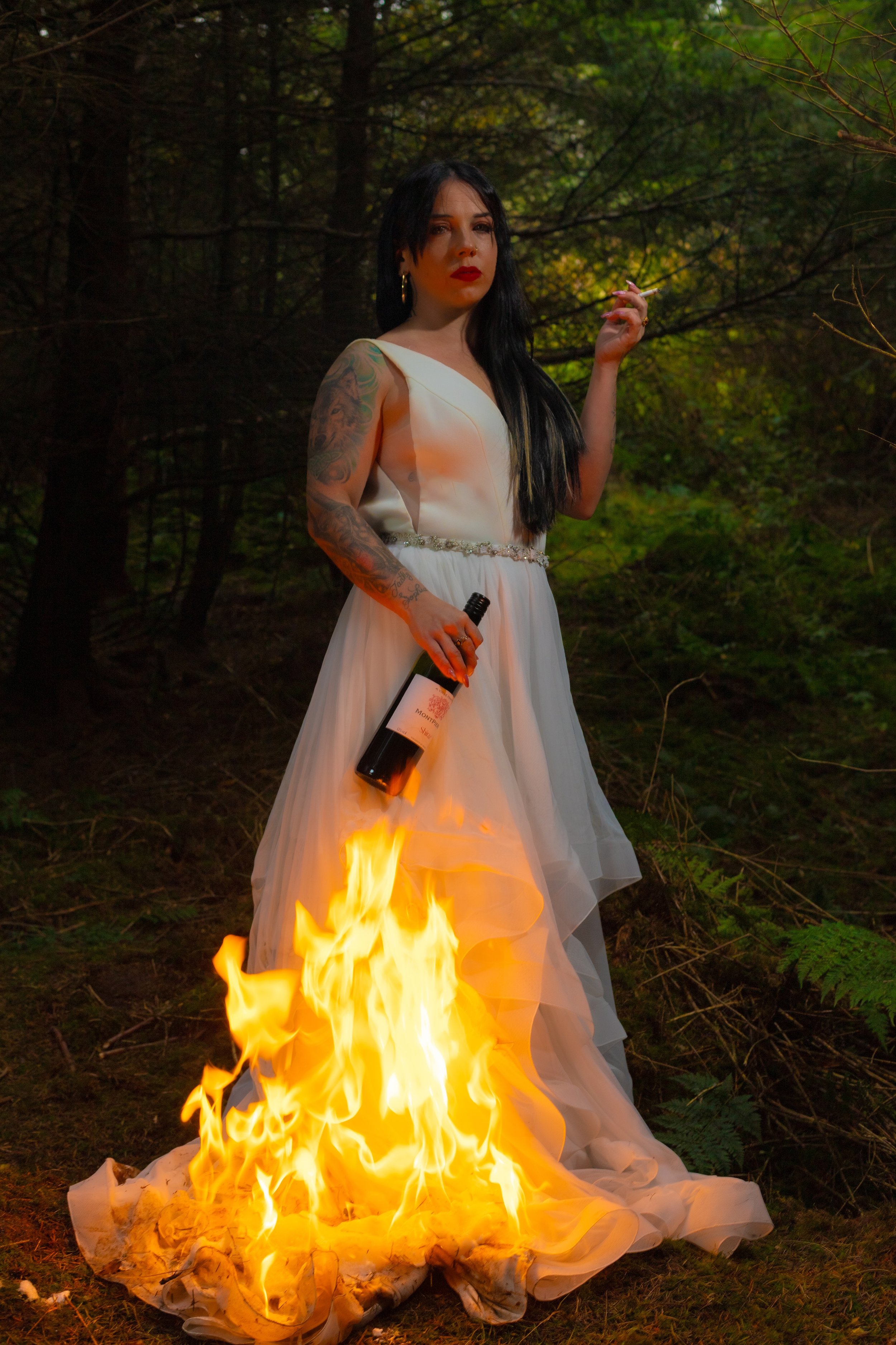 A woman in a white dress stands in a forest with a fire burning beneath her dress, holding a bottle of wine and a cigarette.
