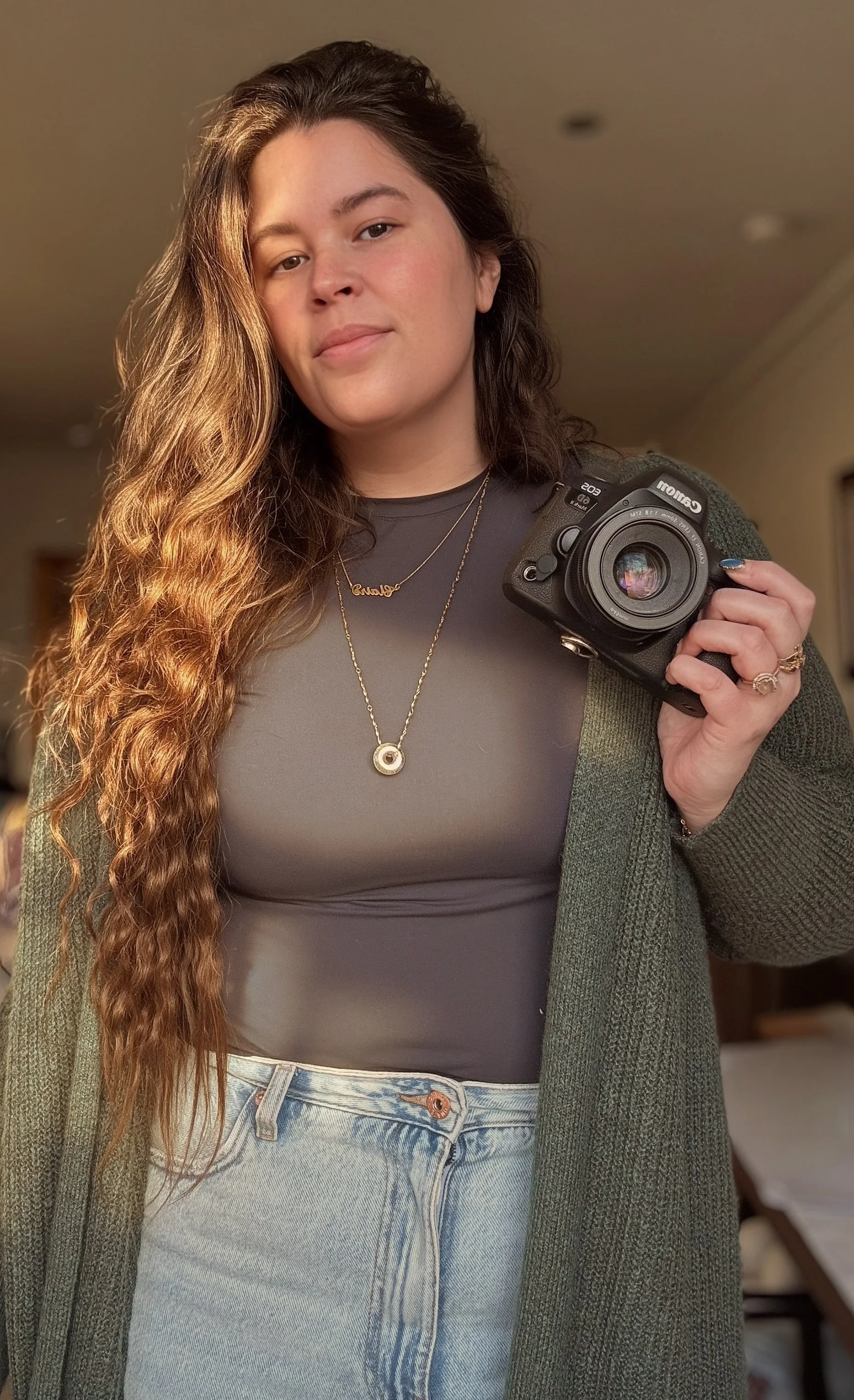 A woman with long, wavy brown hair takes a mirror selfie with a camera in a casual indoor setting. She wears a black top, jeans, layered gold necklaces, and a dark green cardigan.