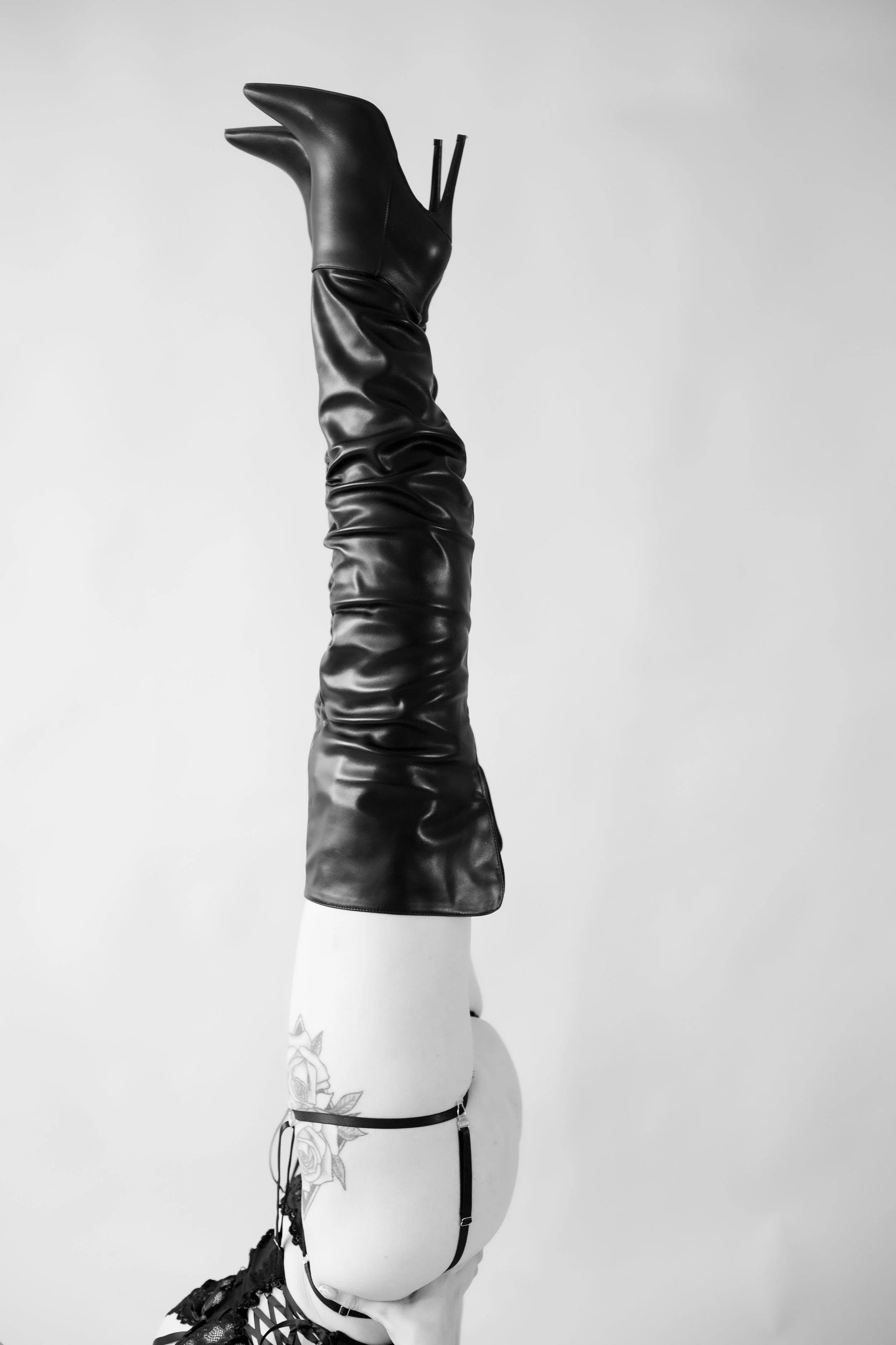 Person in black thigh-high boots and lingerie, performing a handstand with legs extended upward against a plain background.