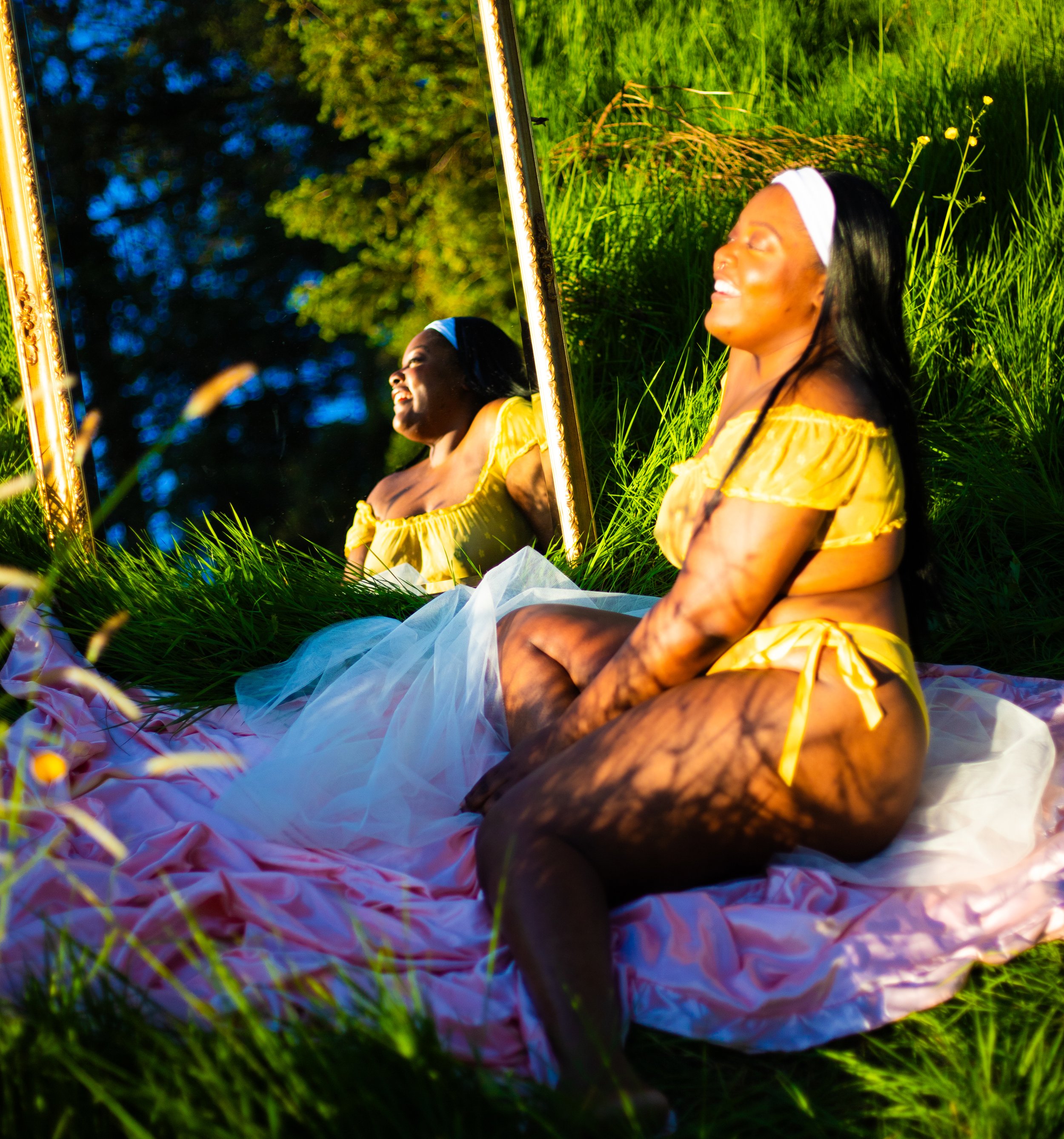 A woman in a yellow off-shoulder top and matching shorts sitting on a pink blanket on grass, looking at her reflection in a mirror outdoors, with sunlight and green trees in the background.