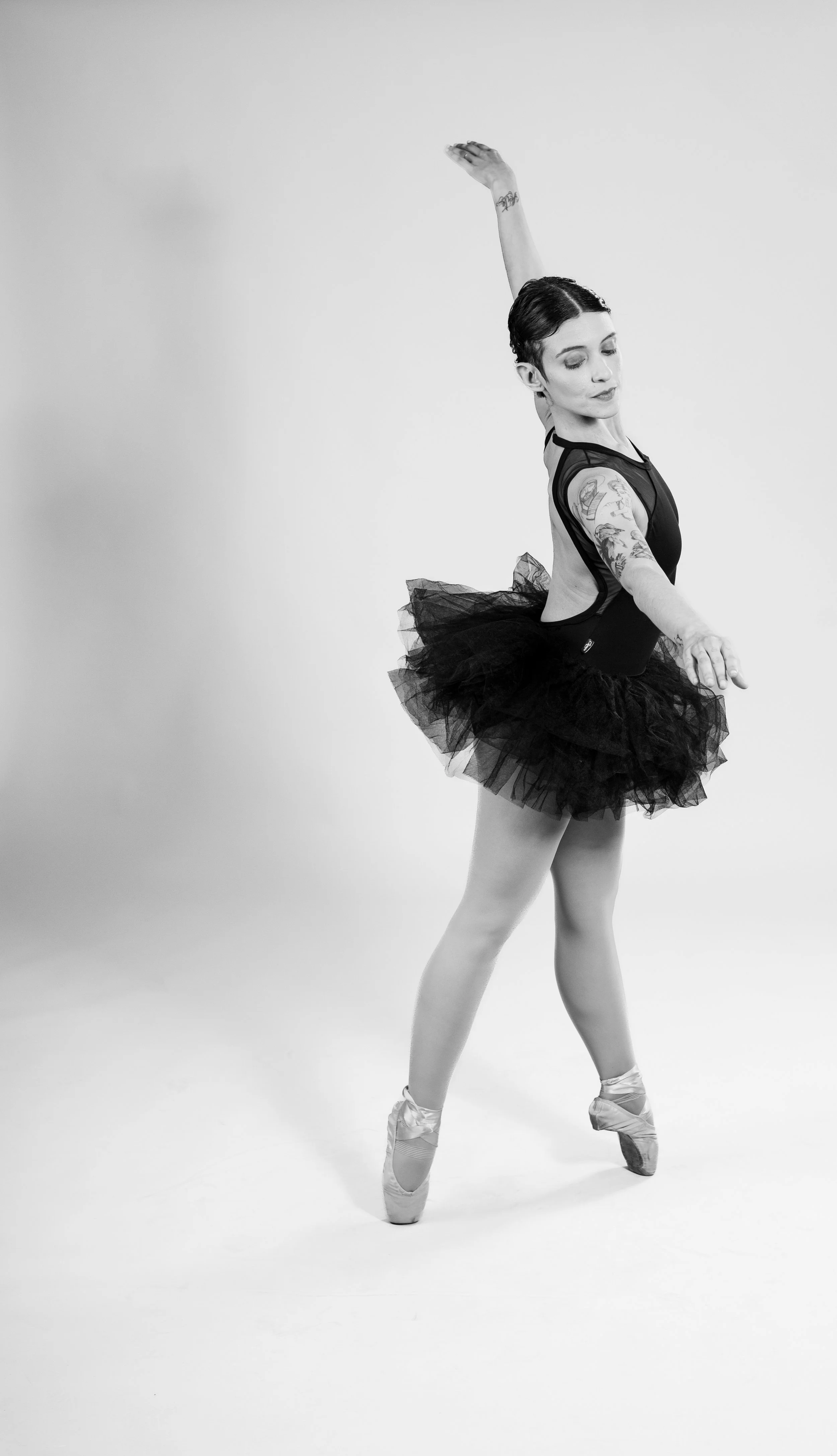 A ballerina in a black tutu and ballet shoes poses with one arm raised and the other extended to the side in a studio setting.