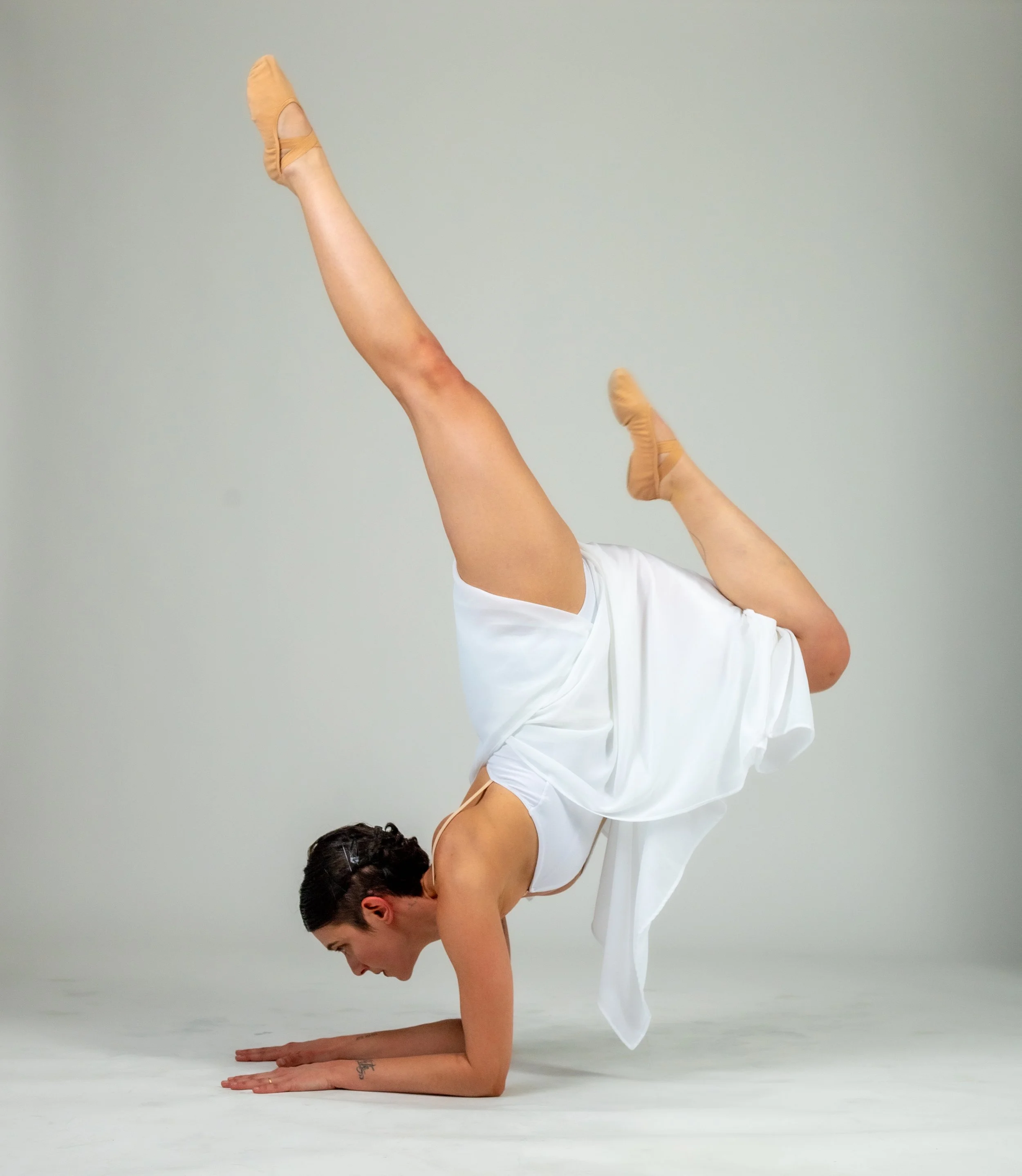 A woman in a white dress performing a handstand with one leg extended upwards and the other bent, on a plain white background.