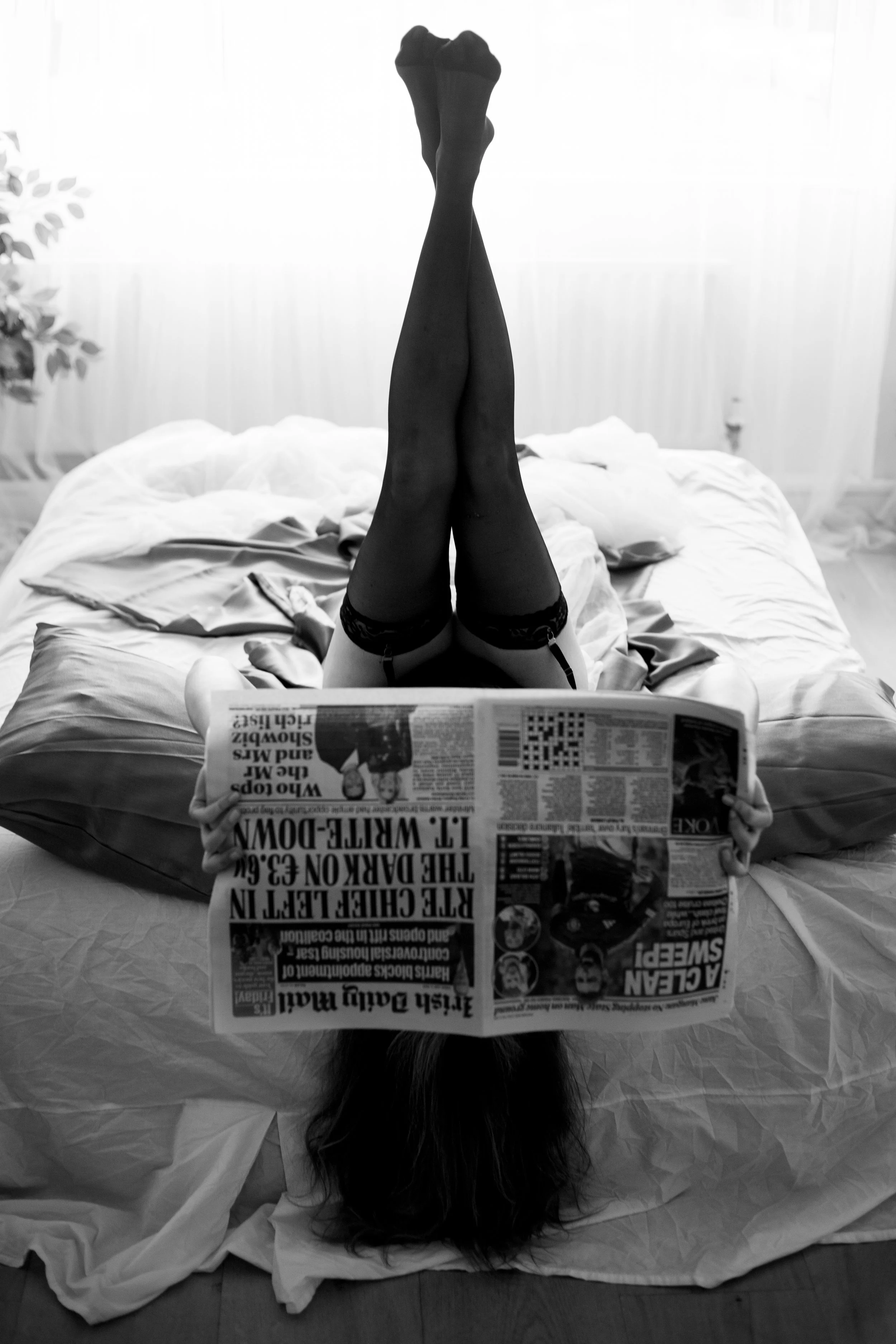 A woman lying upside down on a bed with her legs raised in the air, reading a newspaper. She is wearing thigh-high stockings and the room appears to be a bedroom with a bed, pillows, and a plant in the background.