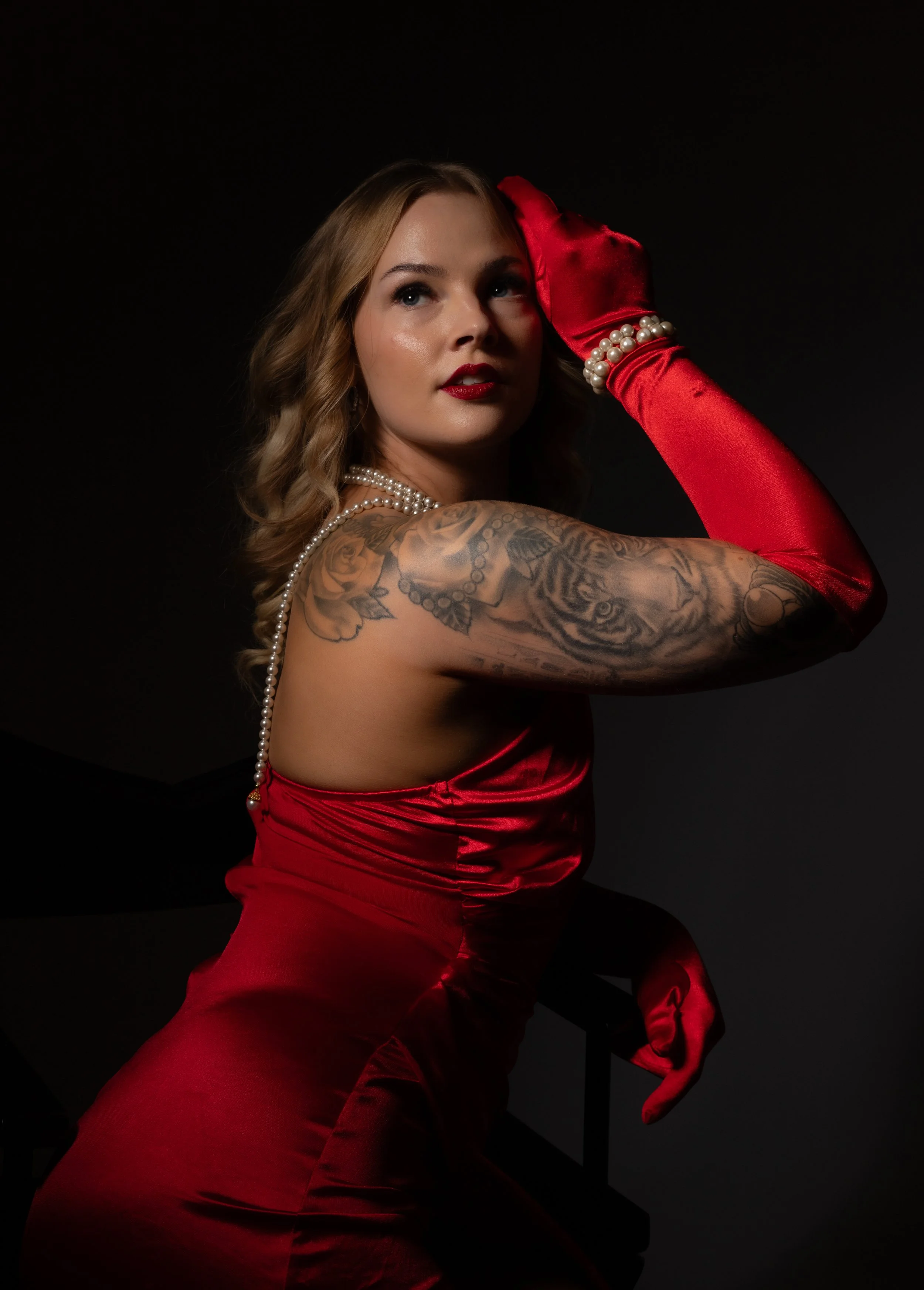A woman with blonde wavy hair and tattoos on her left arm, dressed in a red satin dress, red gloves, pearl necklace, and bracelet, posed against a dark background with her hand on her head and looking slightly upward.