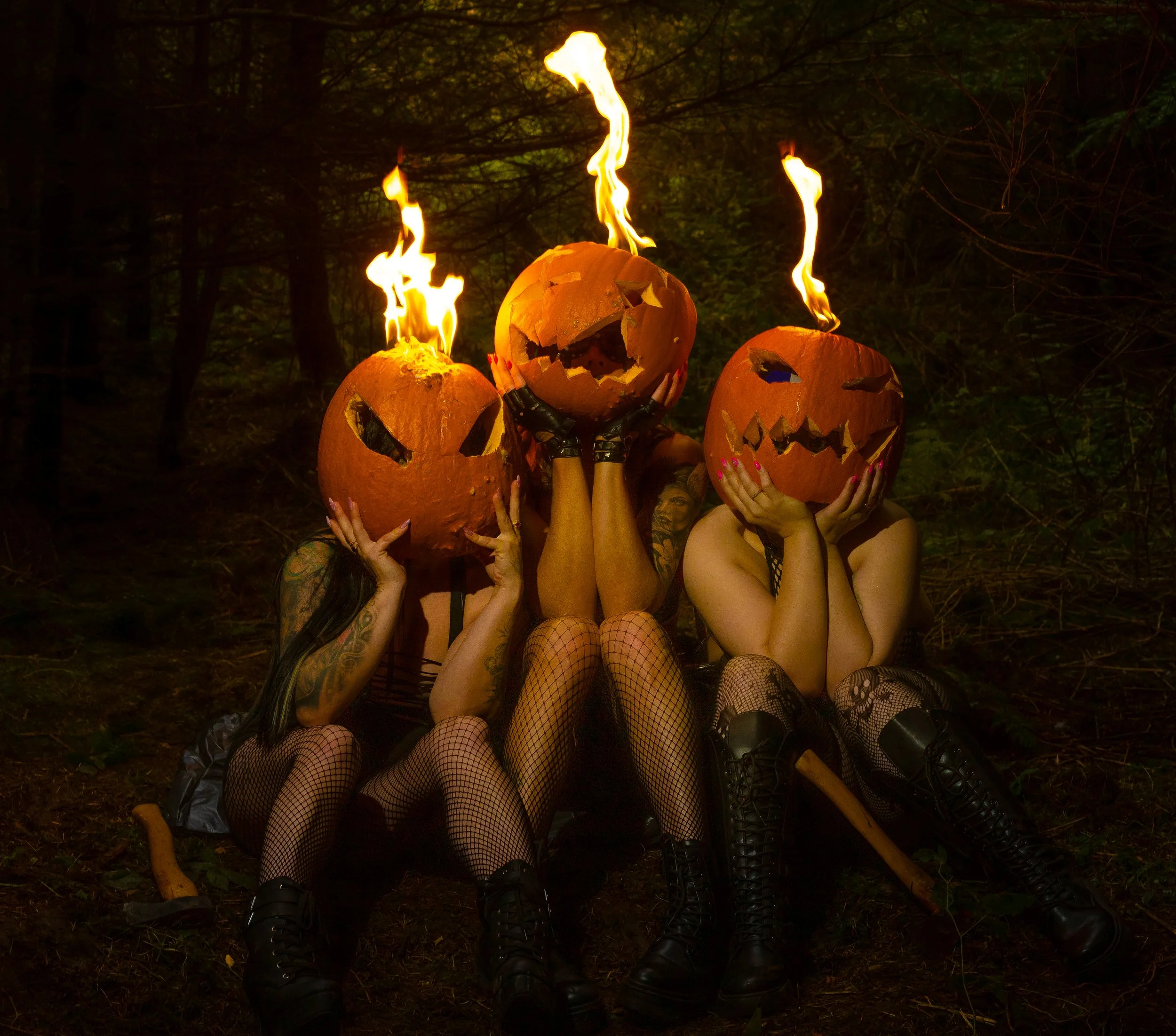 Three women sitting in a dark forest, wearing black outfits with fishnet stockings, each holding a carved pumpkin with a carved face, and flames emerging from the pumpkins' tops.