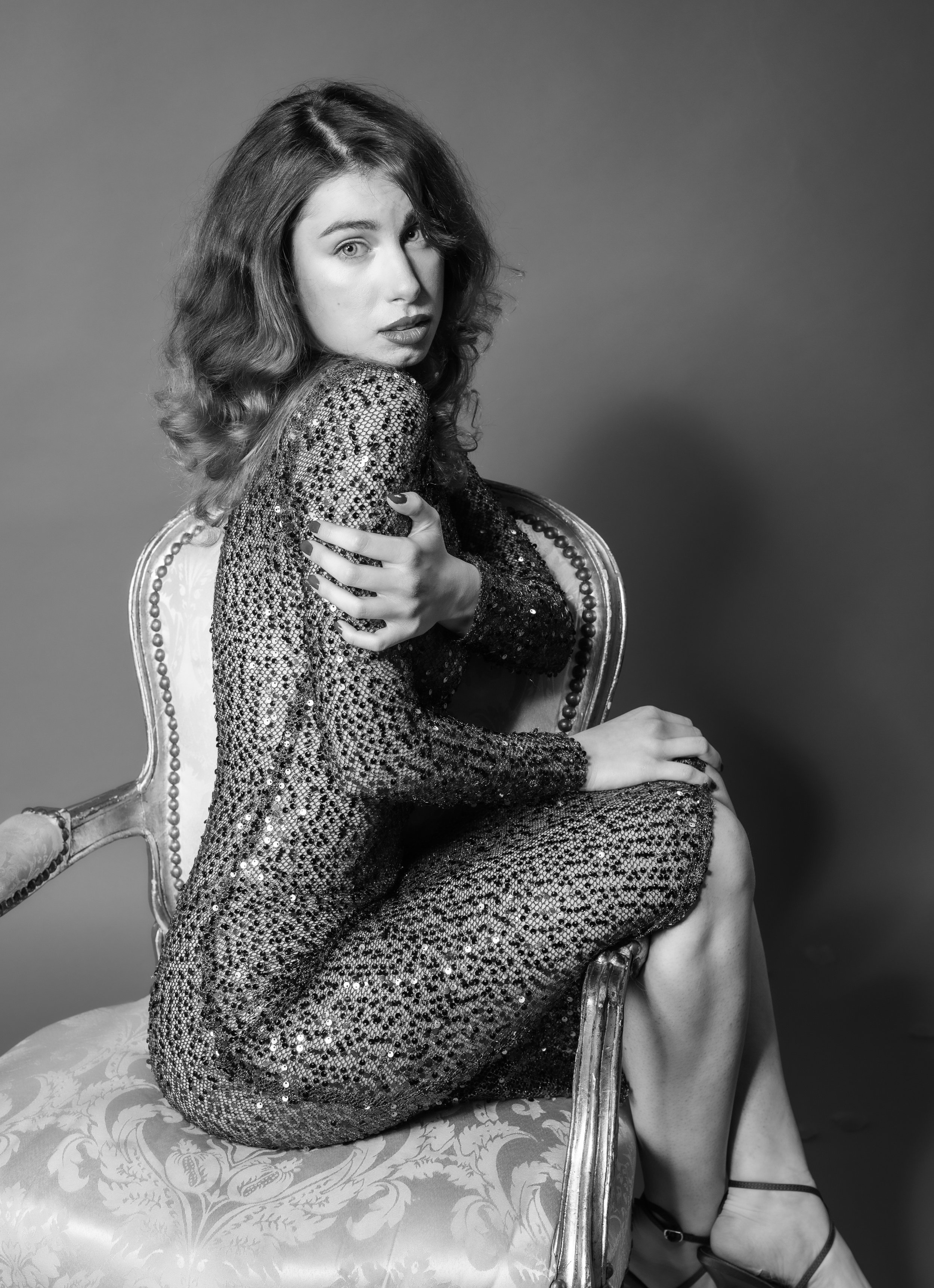 A woman with wavy hair sitting on an ornate chair, wearing a sequined dress, looking at the camera with a serious expression, in black and white.