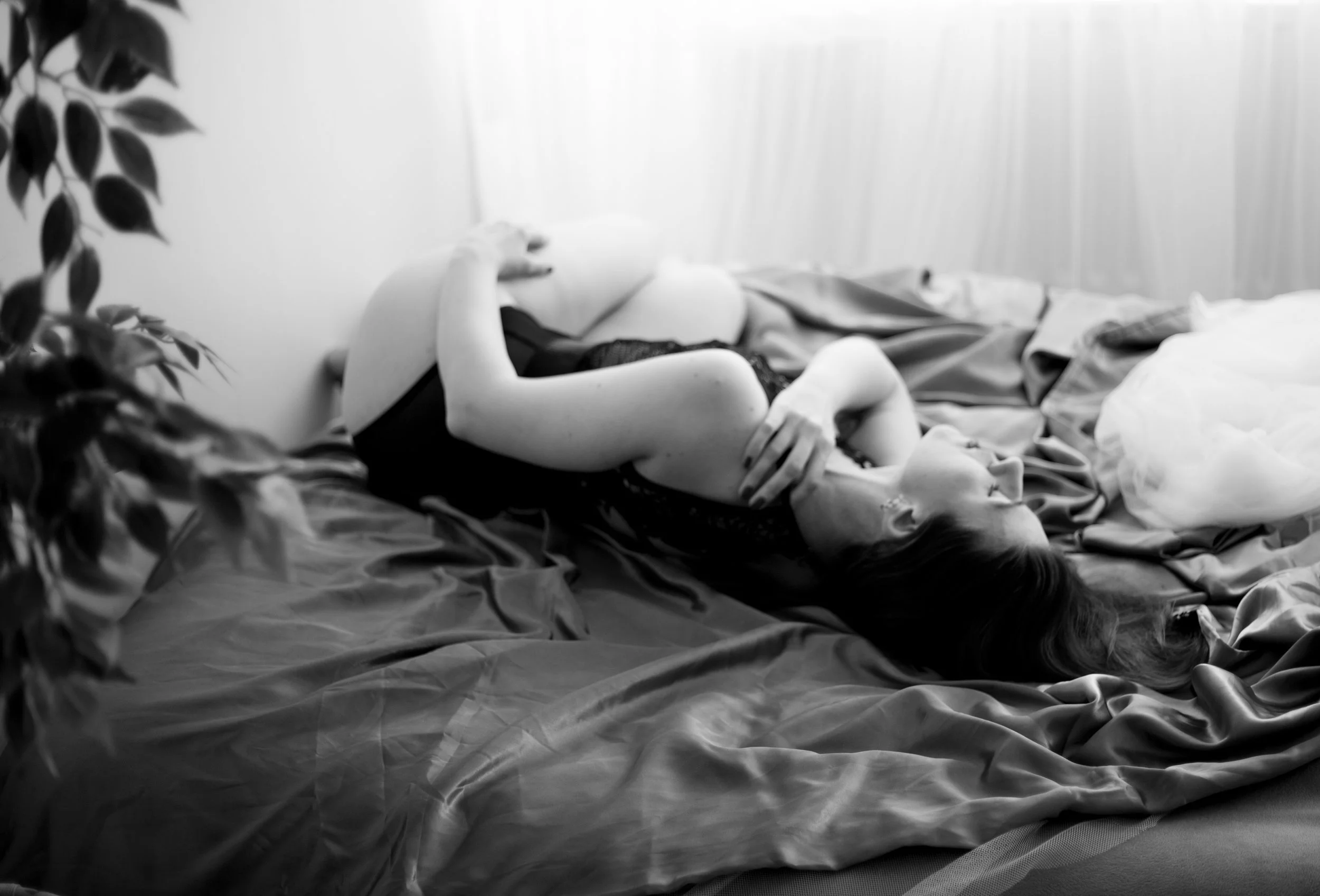 A woman lying on a bed, curled up on her side with her hand on her face, dressed in a black lace outfit, with a bed covered in rumpled sheets and a plant partially visible on the left side.