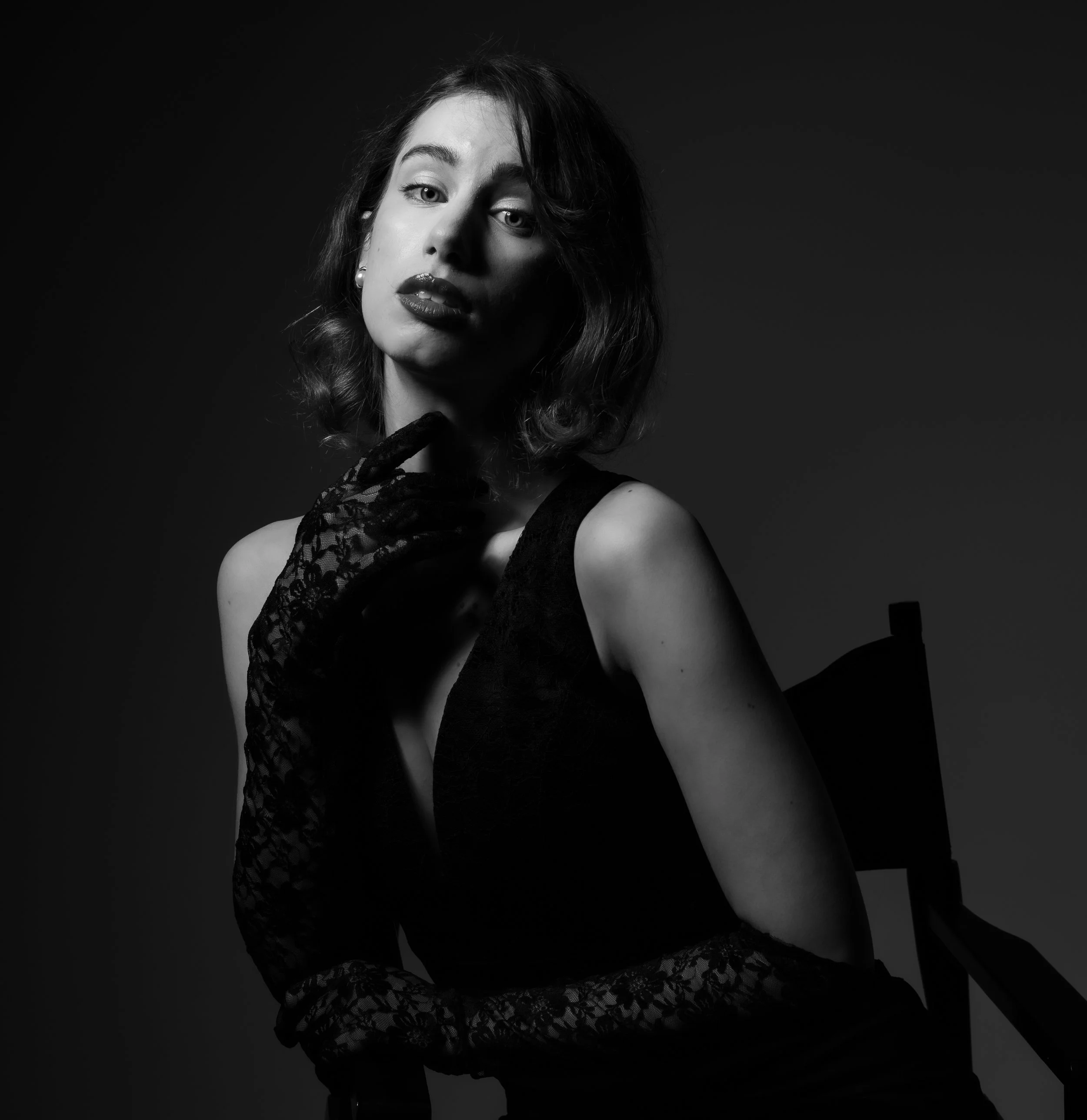A black-and-white portrait of a woman with wavy hair, wearing a sleeveless dress and lace gloves, sitting on a chair with her hand near her chin, against a dark background.