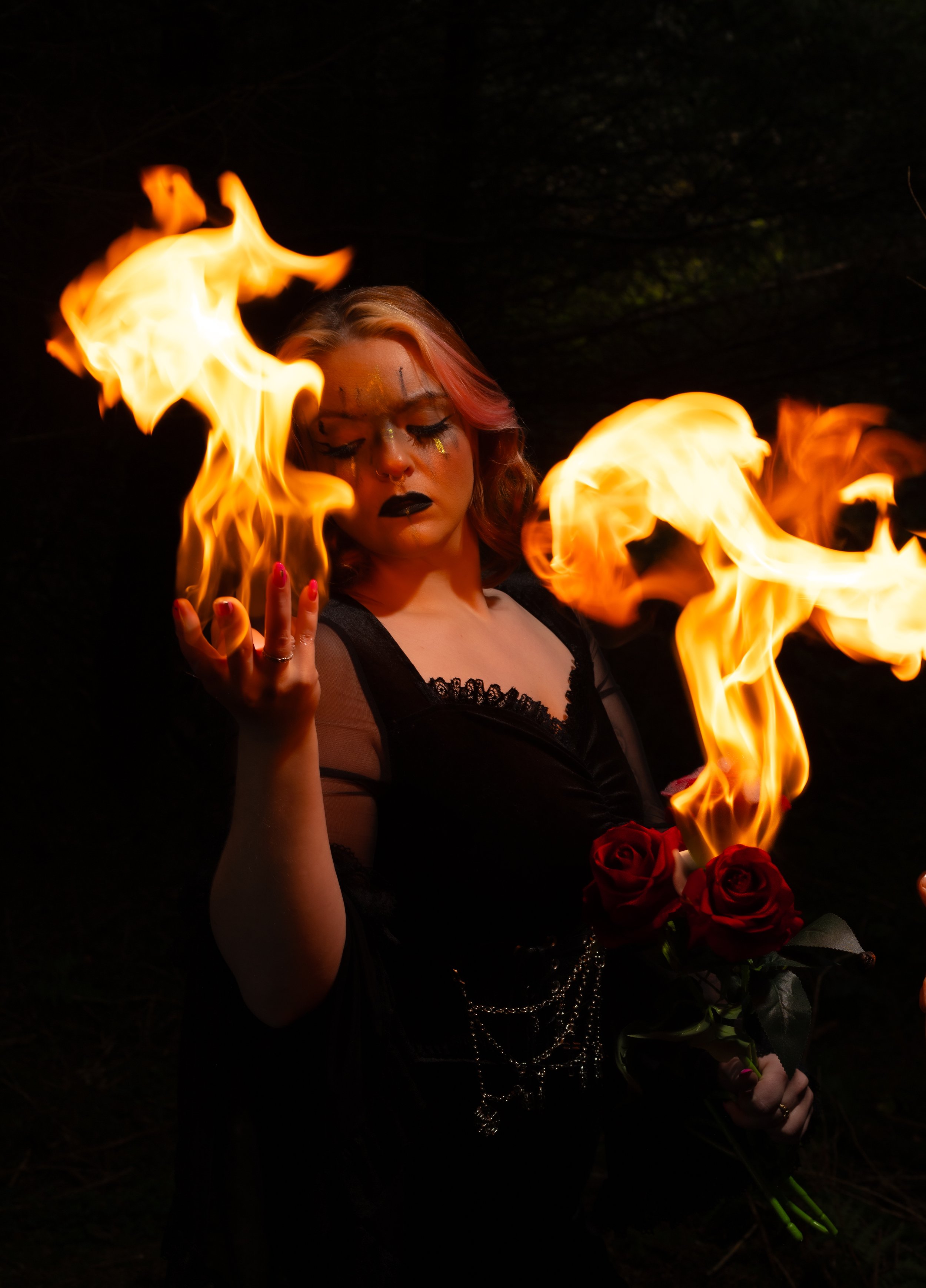 A woman with makeup holding red roses with fire flames surrounding her in a dark setting.