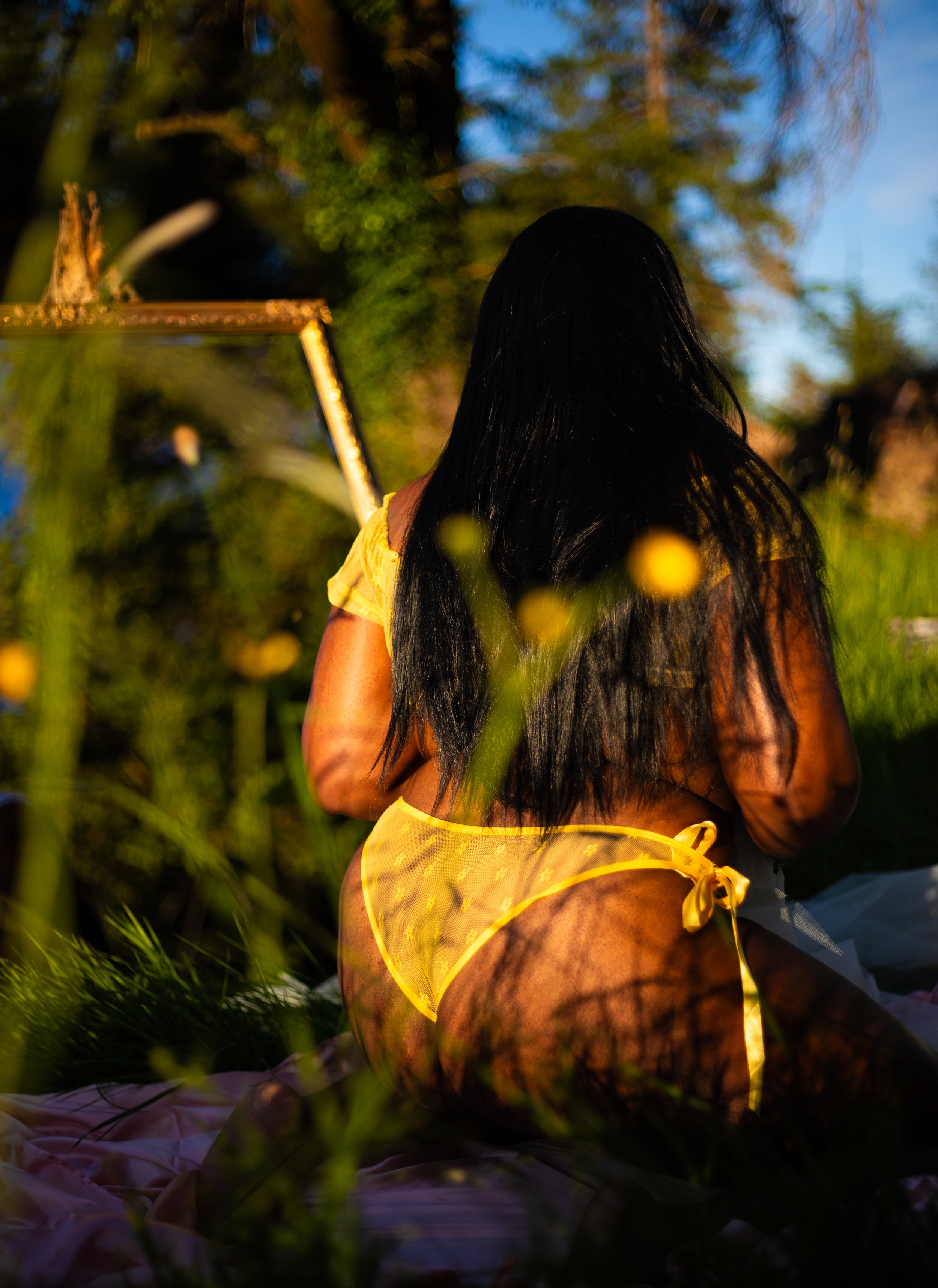 A woman with long black hair wearing yellow lingerie sitting outdoors during sunset, surrounded by greenery and flowers.