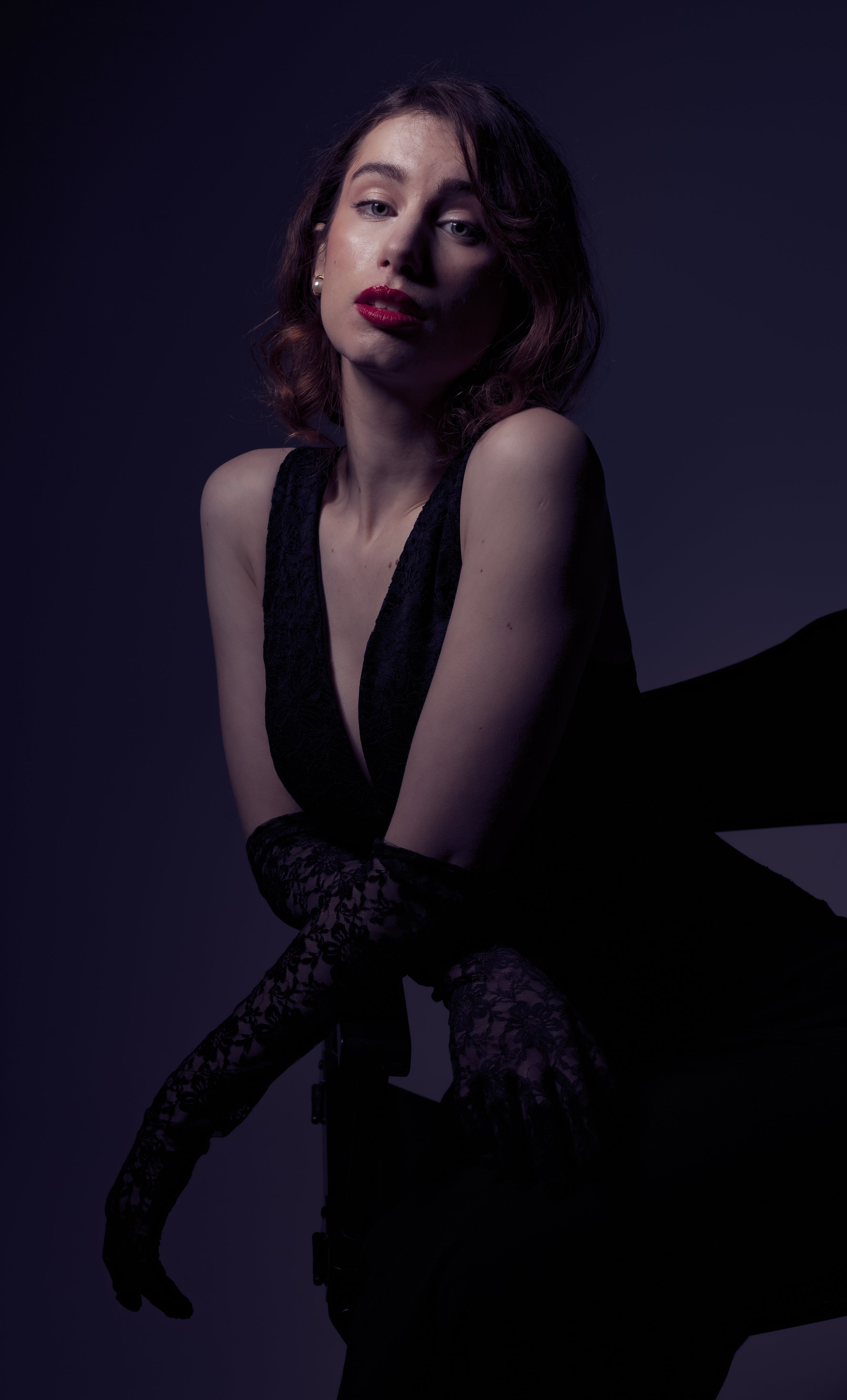 A woman with short, wavy brown hair wearing a black dress and dark lace gloves poses against a dark background with dramatic lighting.