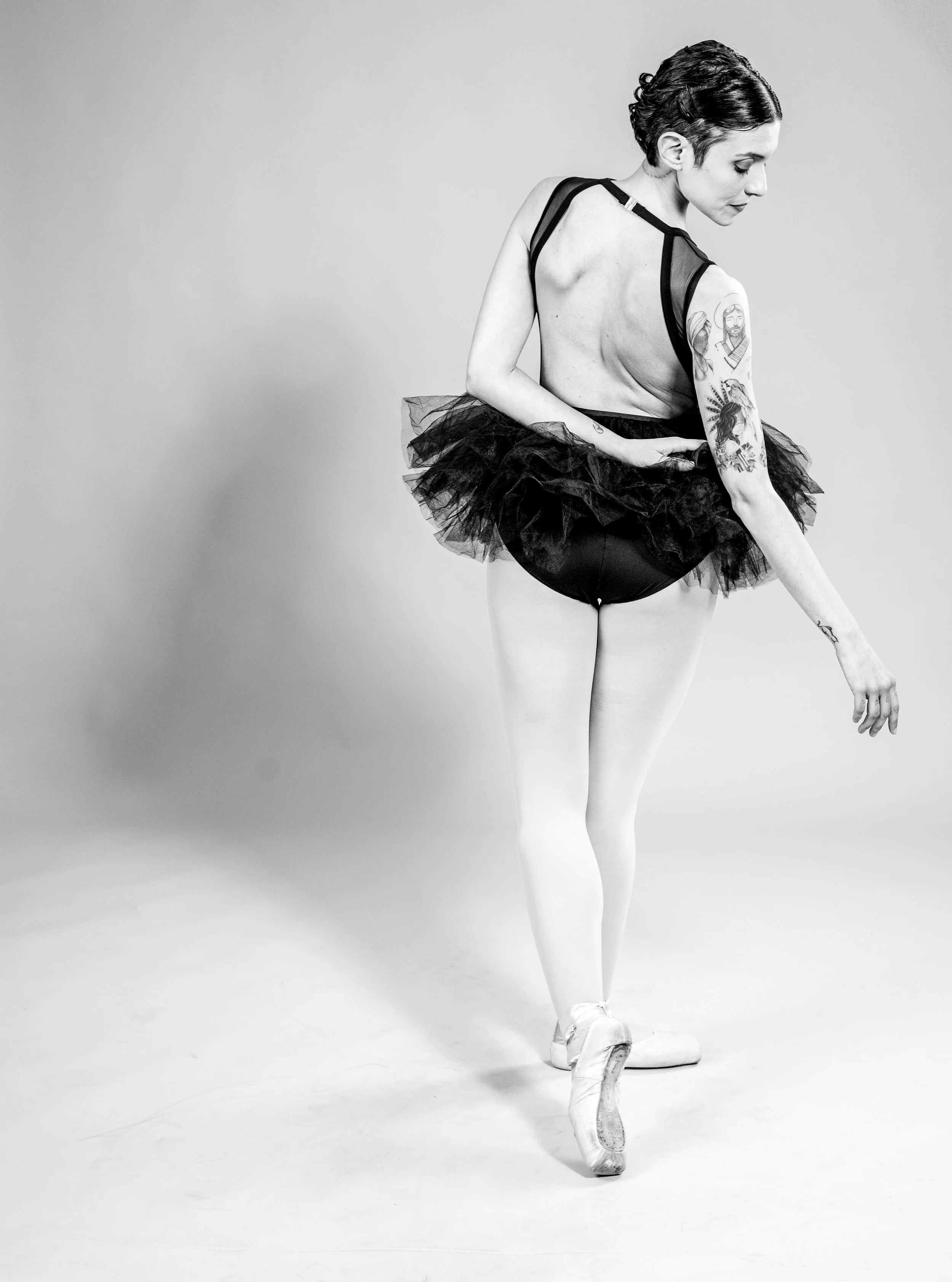 A woman dressed as a ballerina with a tutu, wearing ballet shoes, standing on one foot and pointing with her right hand.