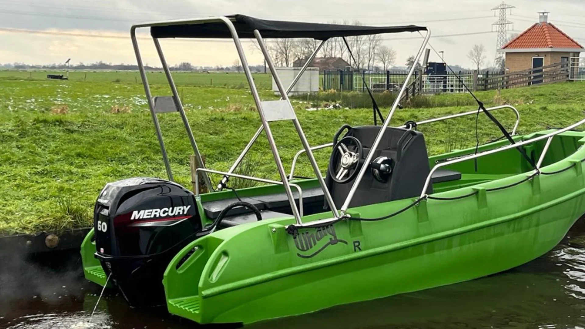 A green boat with a Mercury 60 horsepower outboard motor docked beside a grassy shoreline, with open fields, a fence, and a building with a red roof in the background.