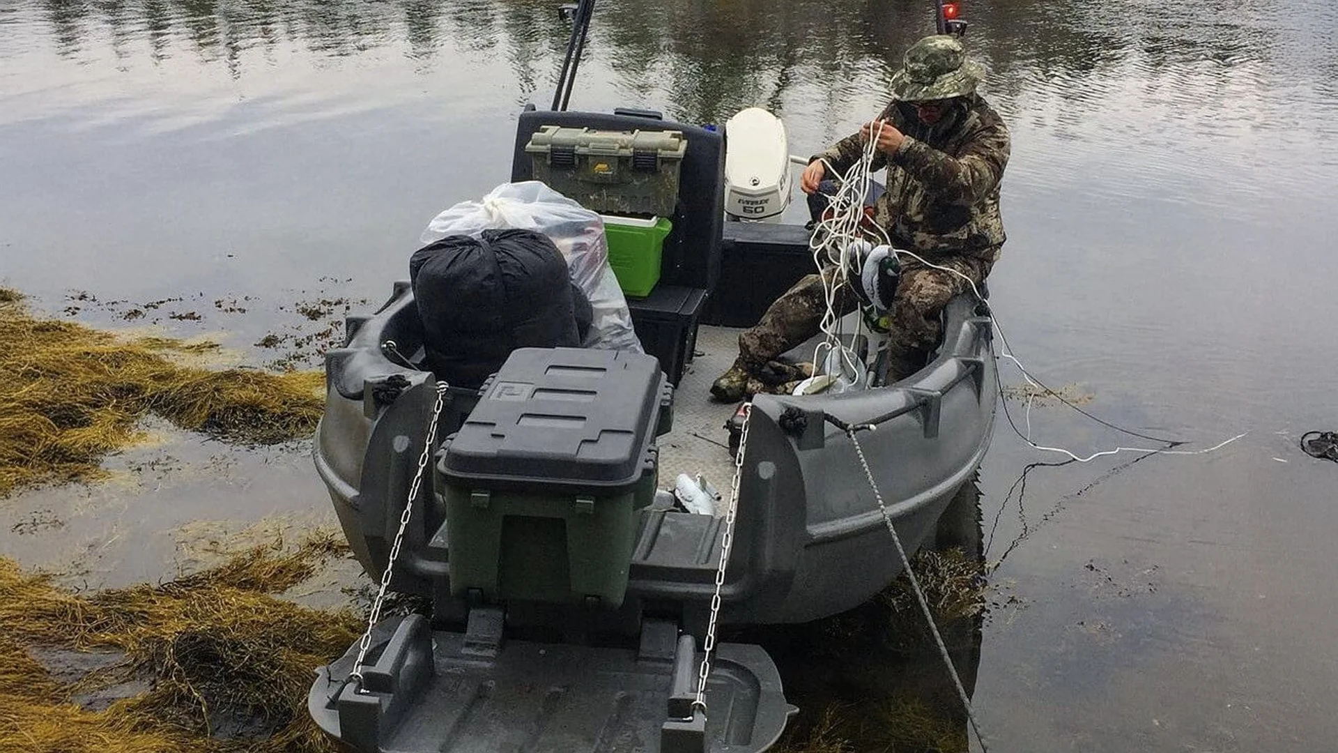 A person in camouflage clothing and a hat sitting in a small gray boat on a body of water, tying or untangling white ropes. The boat is filled with gear, including a green cooler, black and white bags, and tackle boxes, with seaweed along the water's edge.