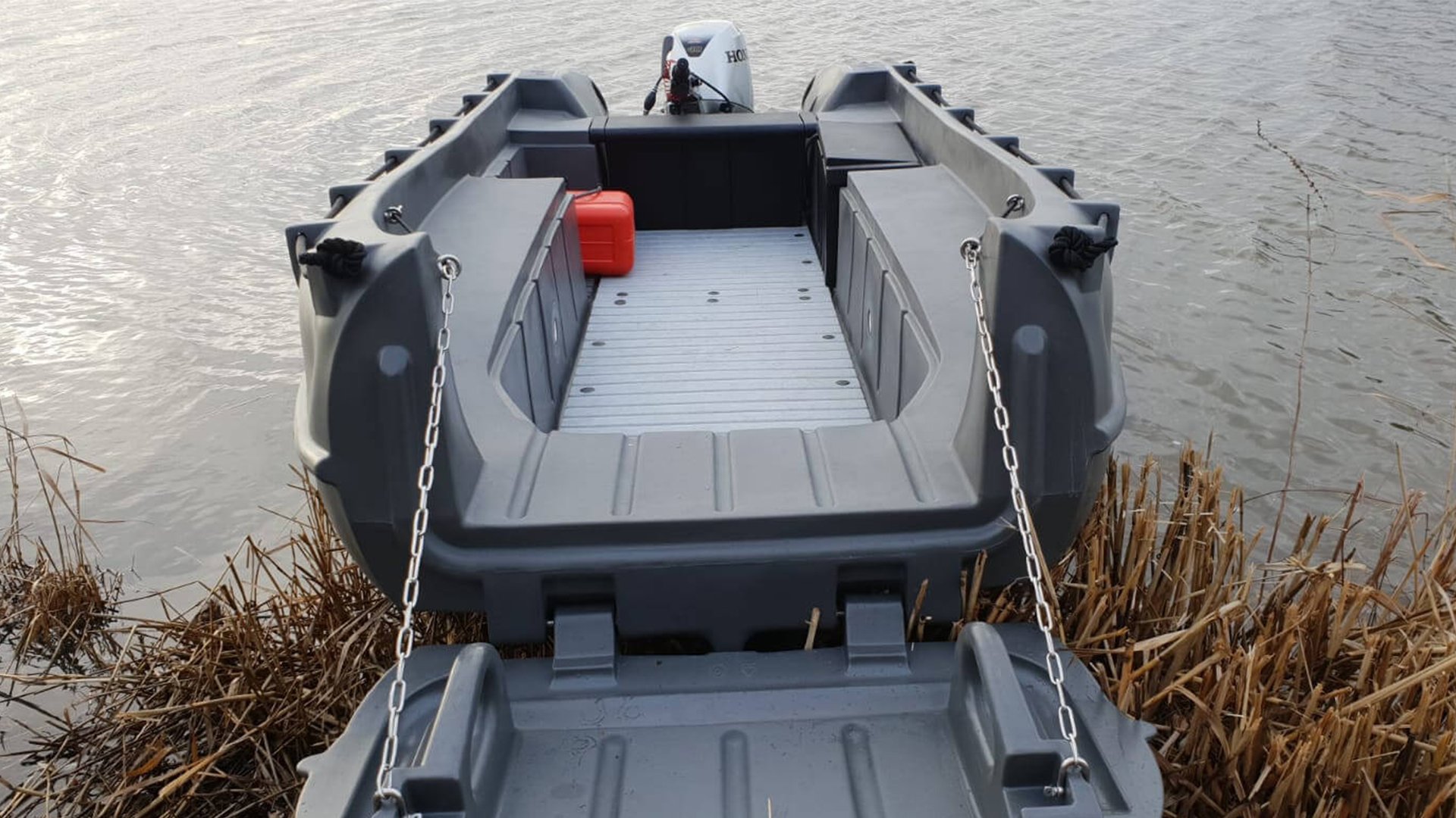 A small gray motorboat docked on a lakeside, with brown reed plants nearby, a mounted Honda outboard motor, a red fuel container inside, and chains securing the boat to the shore.