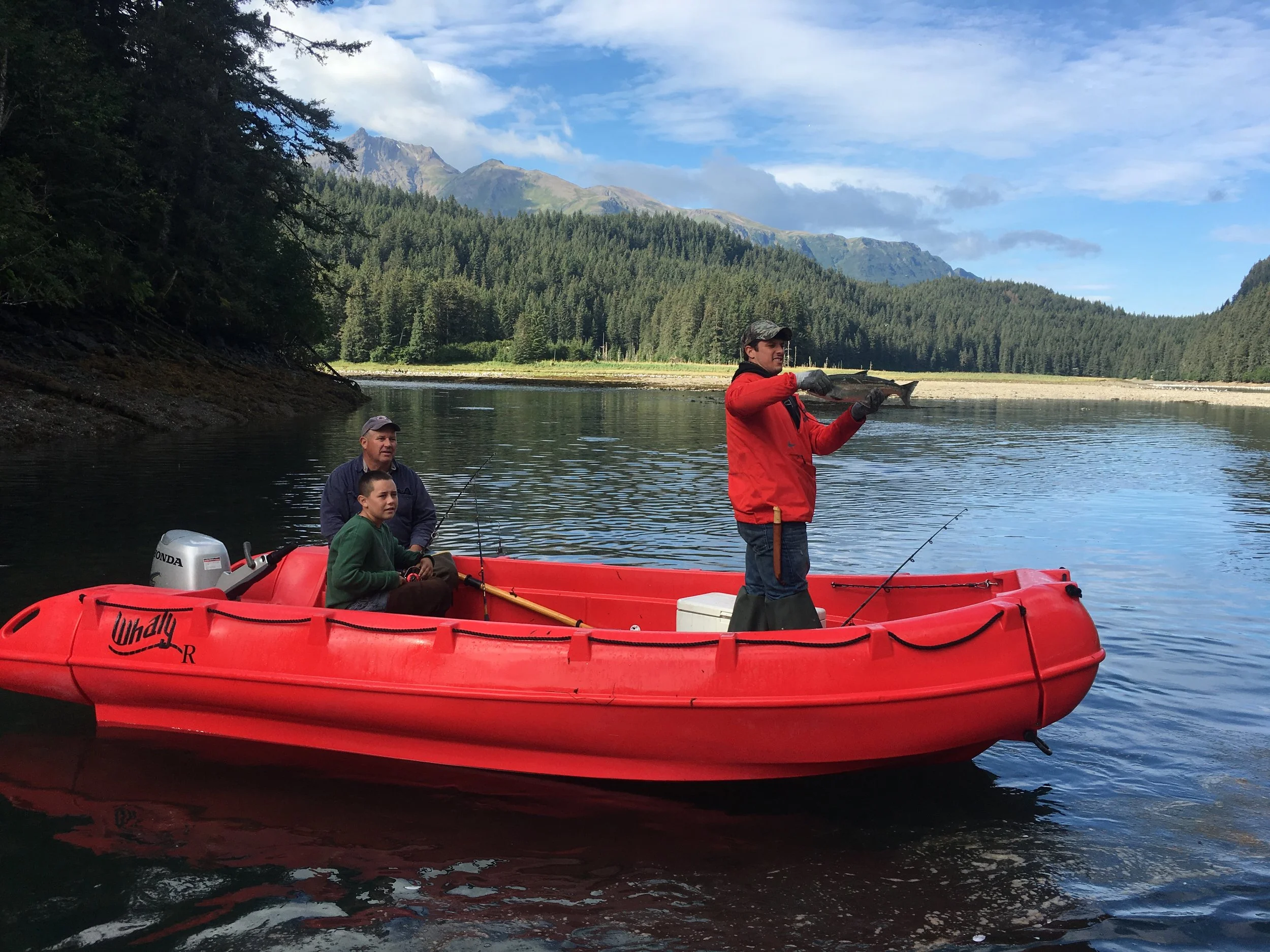 Three people on a red boat on a lake surrounded by trees and mountains. One person is standing and holding a fish, while the other two are sitting with fishing rods.