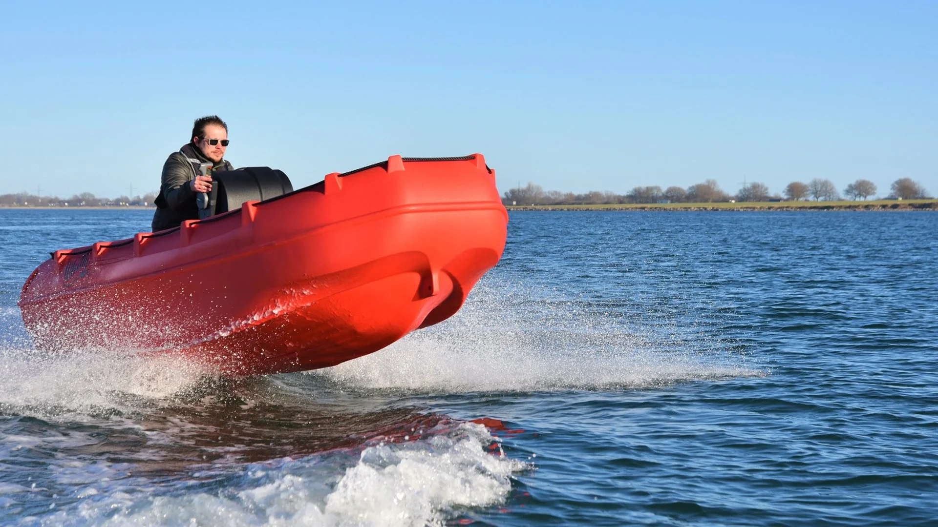 Man in sunglasses navigating a red motorboat on a calm body of water with a distant shoreline and clear blue sky.