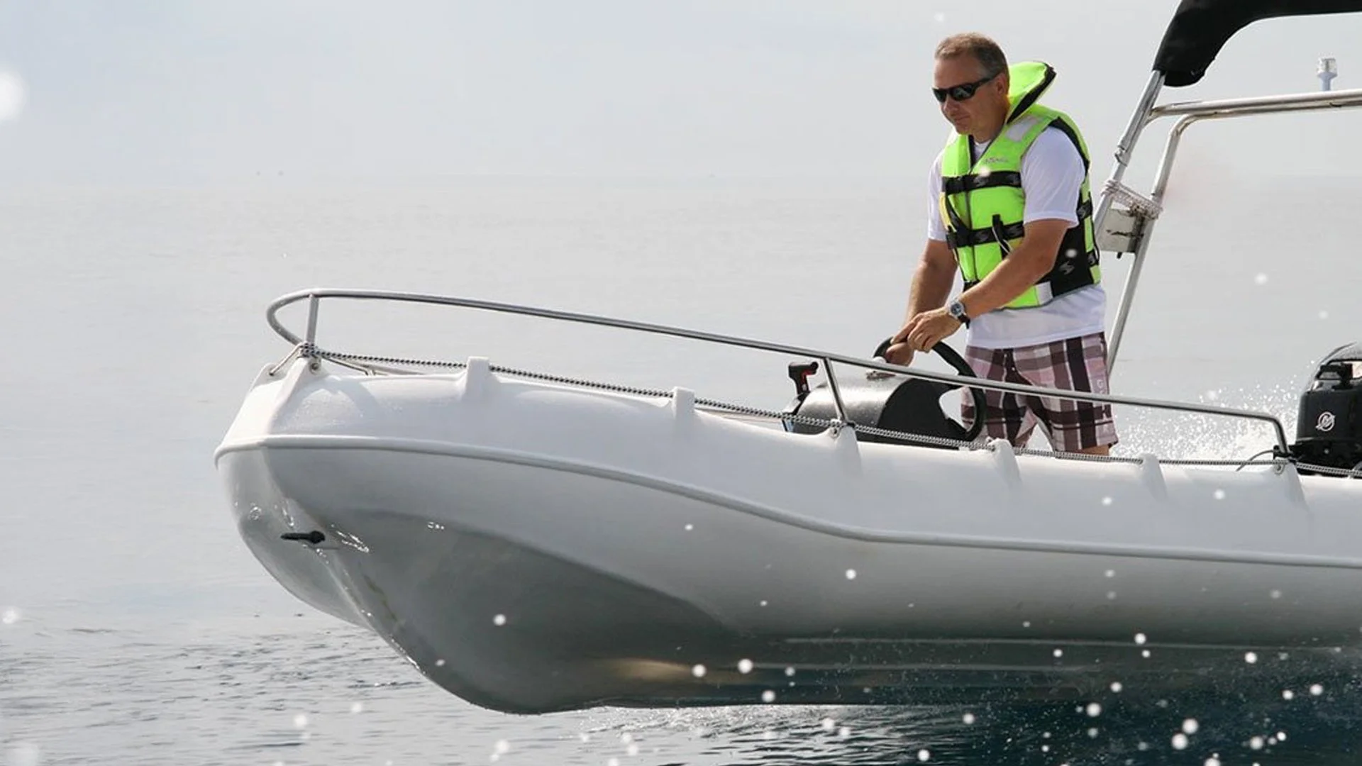 Man wearing sunglasses and a yellow life vest, standing on a small boat, steering with a wheel, dressed in a white T-shirt and plaid shorts, on a body of water.