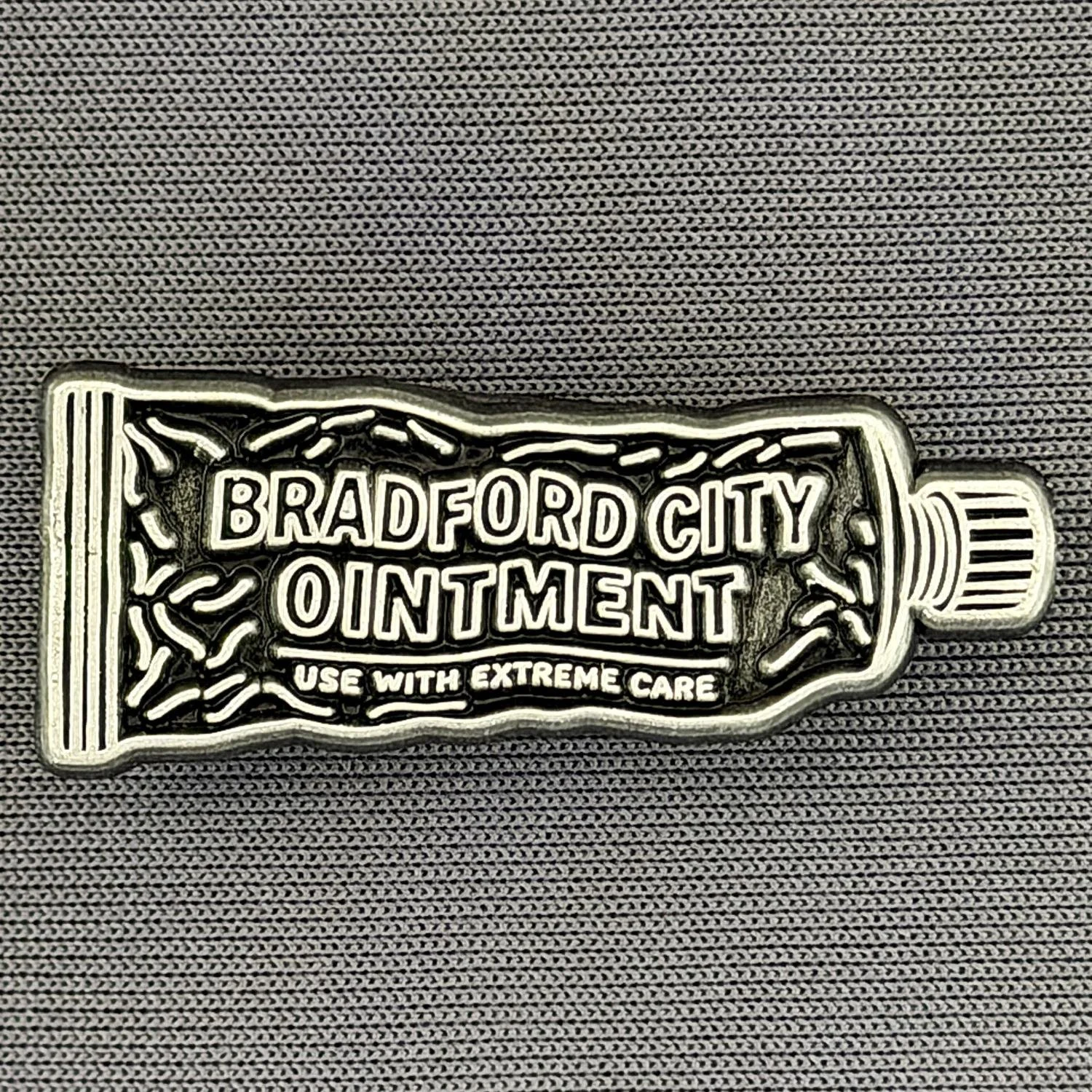 A metal badge shaped like a tube of ointment labeled 'Bradford City Ointment' with the words 'Use with extreme care' below, placed on a textured gray fabric background.