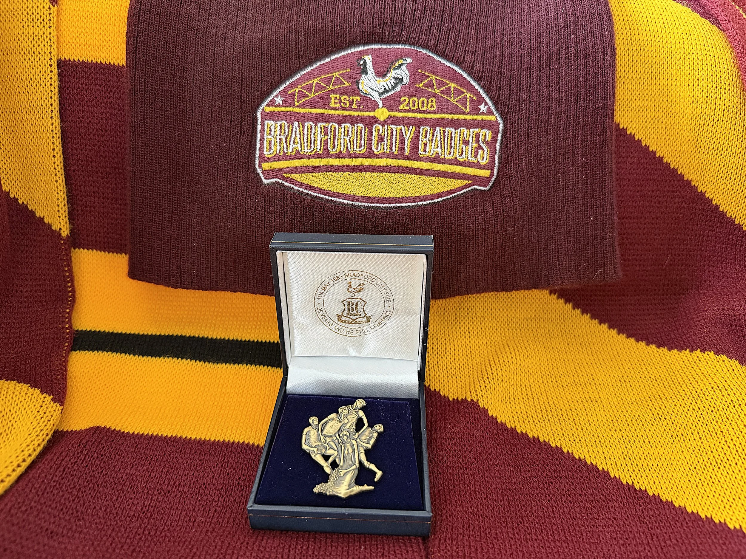 Close-up view of a maroon and yellow sports jersey with an embroidered badge that reads 'Bradford City Badges, Est. 2008' and a crest featuring a griffin. Below the badge, there is a medal with a relief image of a football scene.