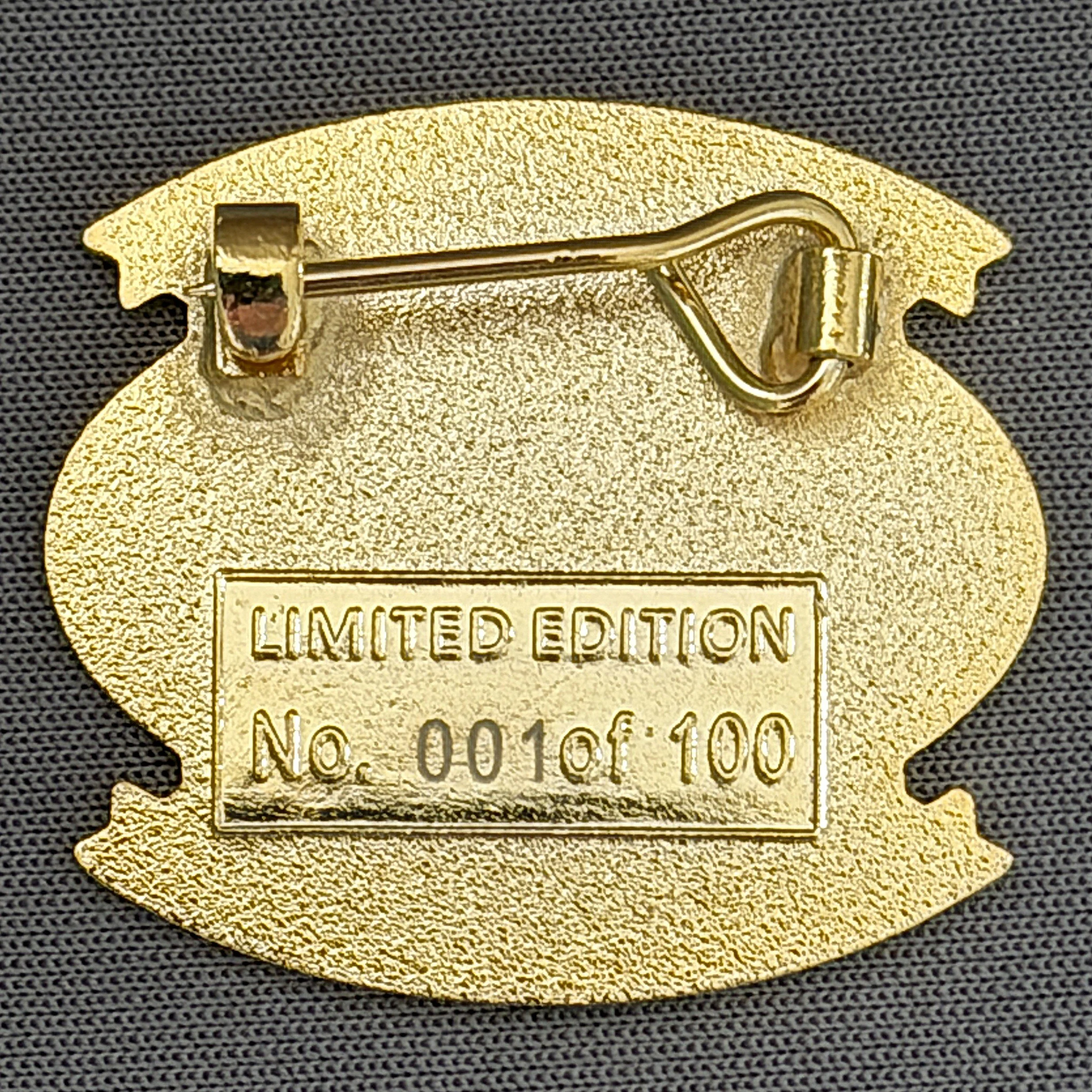 Gold-colored pin with a safety pin design and the words 'Limited Edition' engraved on it, attached to a dark fabric background.
