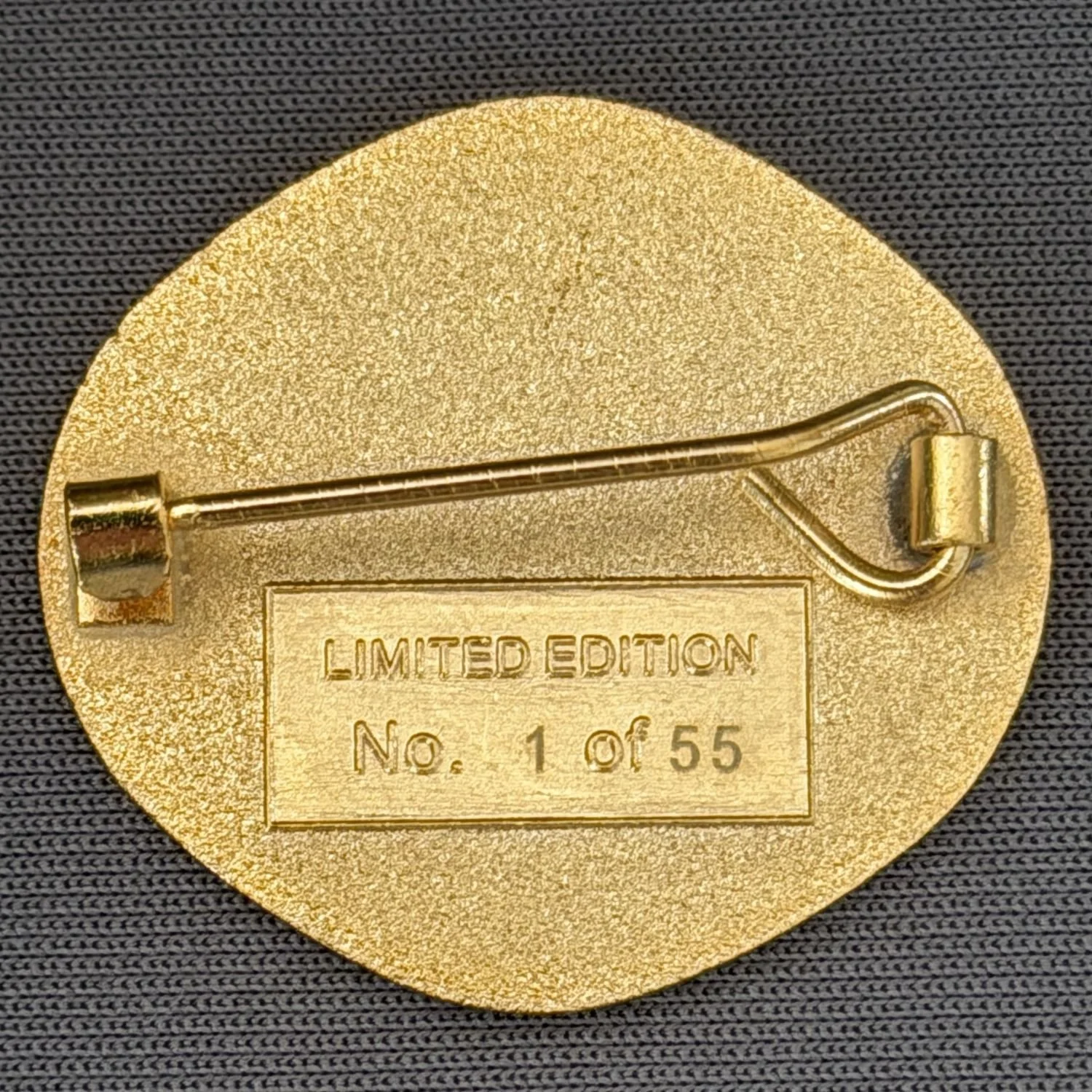 Back of a pin with a safety pin clasp, gold in color, on dark fabric. The pin has a rectangular label that reads 'Limited Edition No. 1 of 55'.