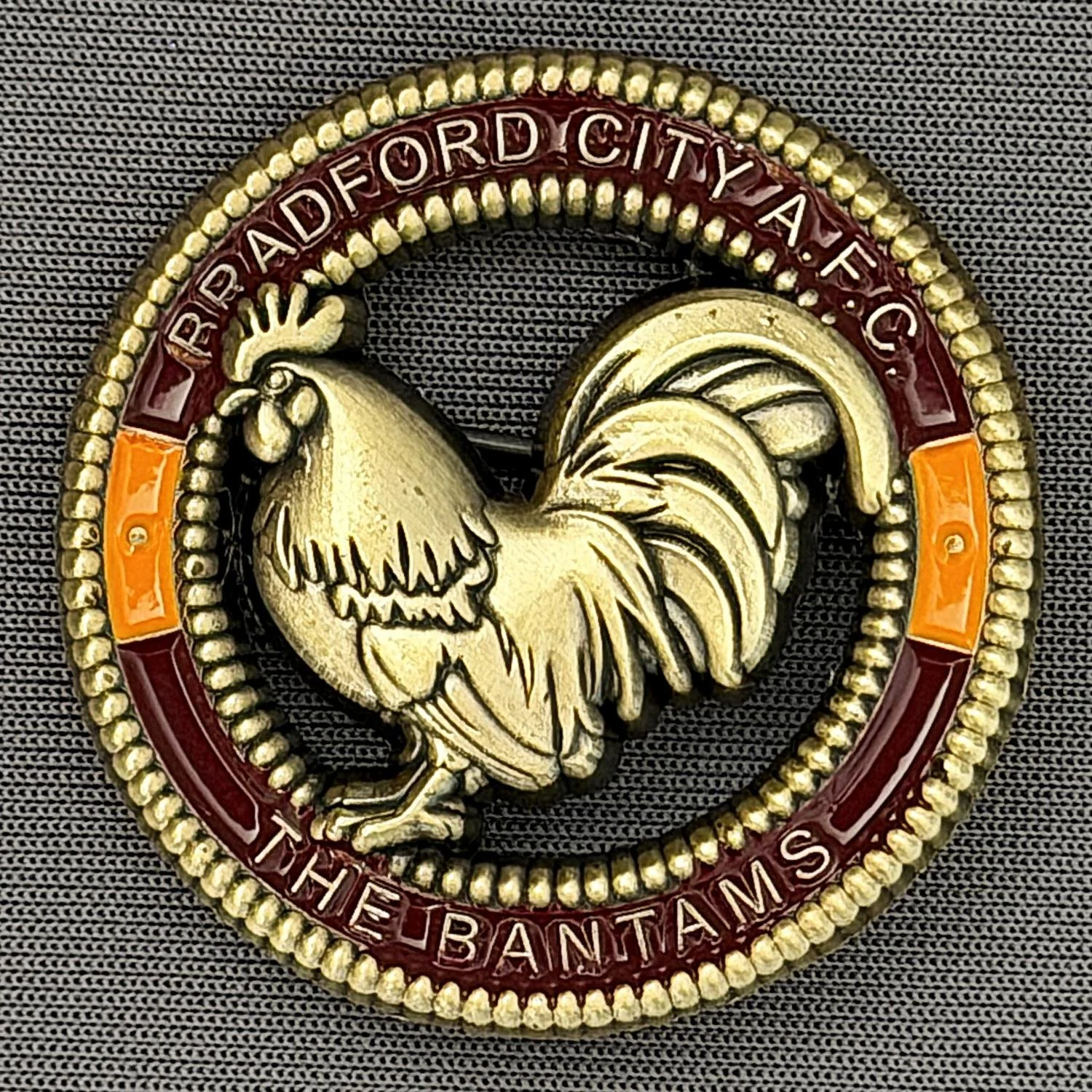 Close-up of a circular badge with a gold-colored rooster in the center, surrounded by a beaded border and an inscription that reads 'City of Ford' and 'The Banta M', with red, orange, and gold colors on a dark textured background.