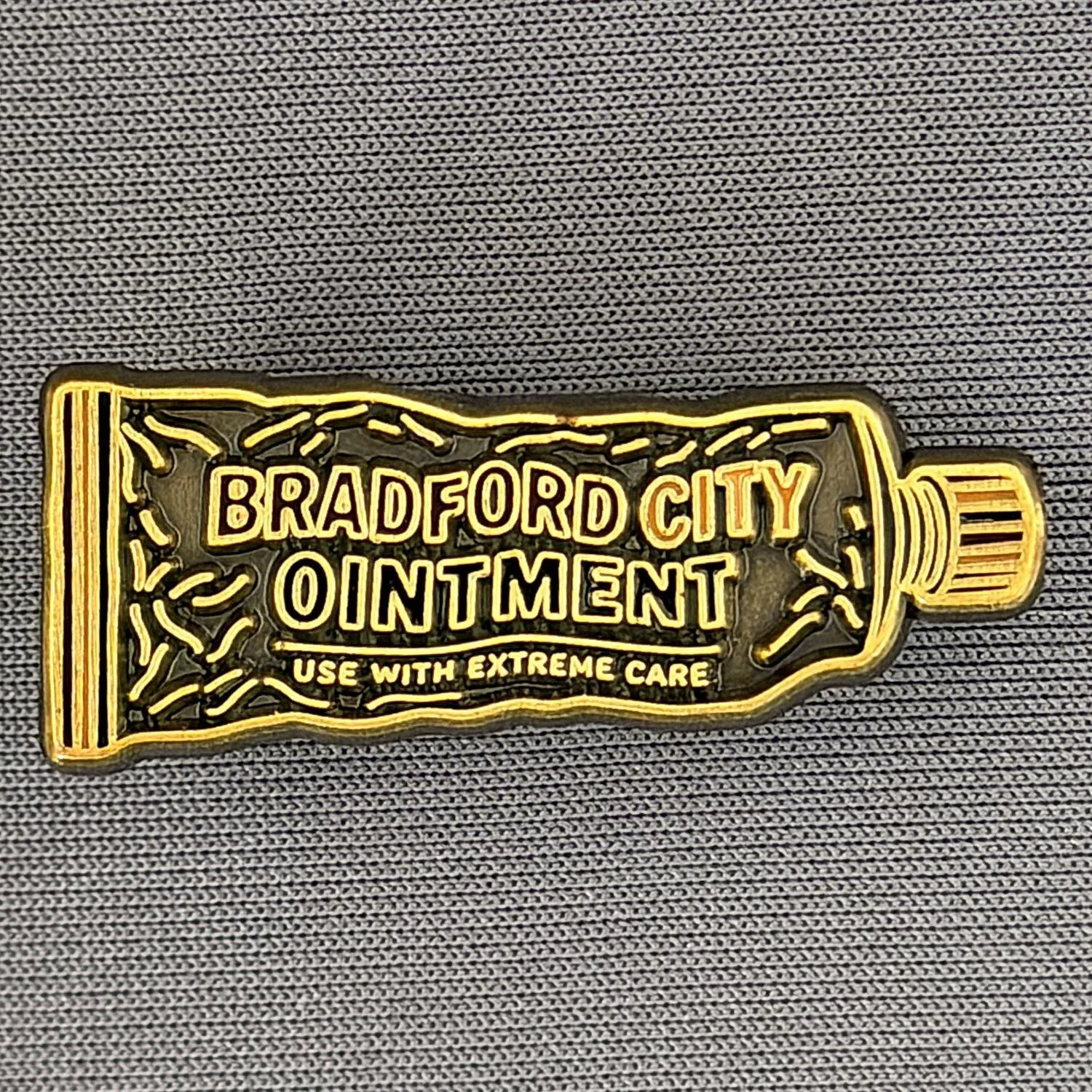 A pin in the shape of a crayon with yellow and black outlines. It features the text "Bradford City Ointment" and a smaller caption "Use with extreme care" on a patterned fabric background.