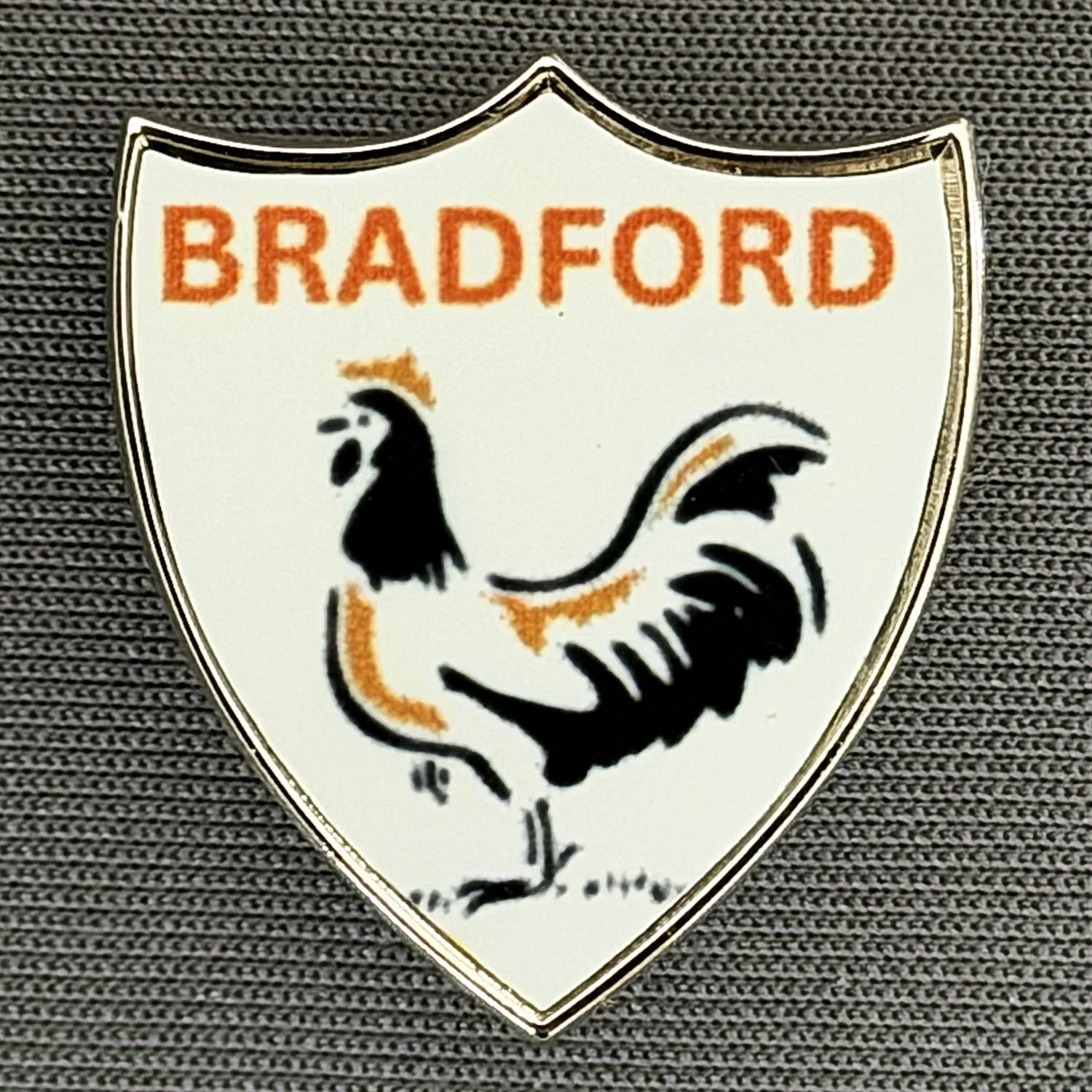 A shield-shaped badge with the word "Bradford" in orange at the top. Below it, a black and orange stylized rooster is depicted. The badge is placed on a dark, textured fabric background.
