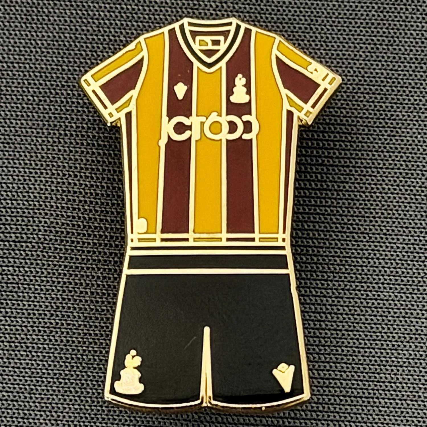 Pin of a soccer jersey with yellow and maroon vertical stripes, black shorts, and small symbols on the jersey.