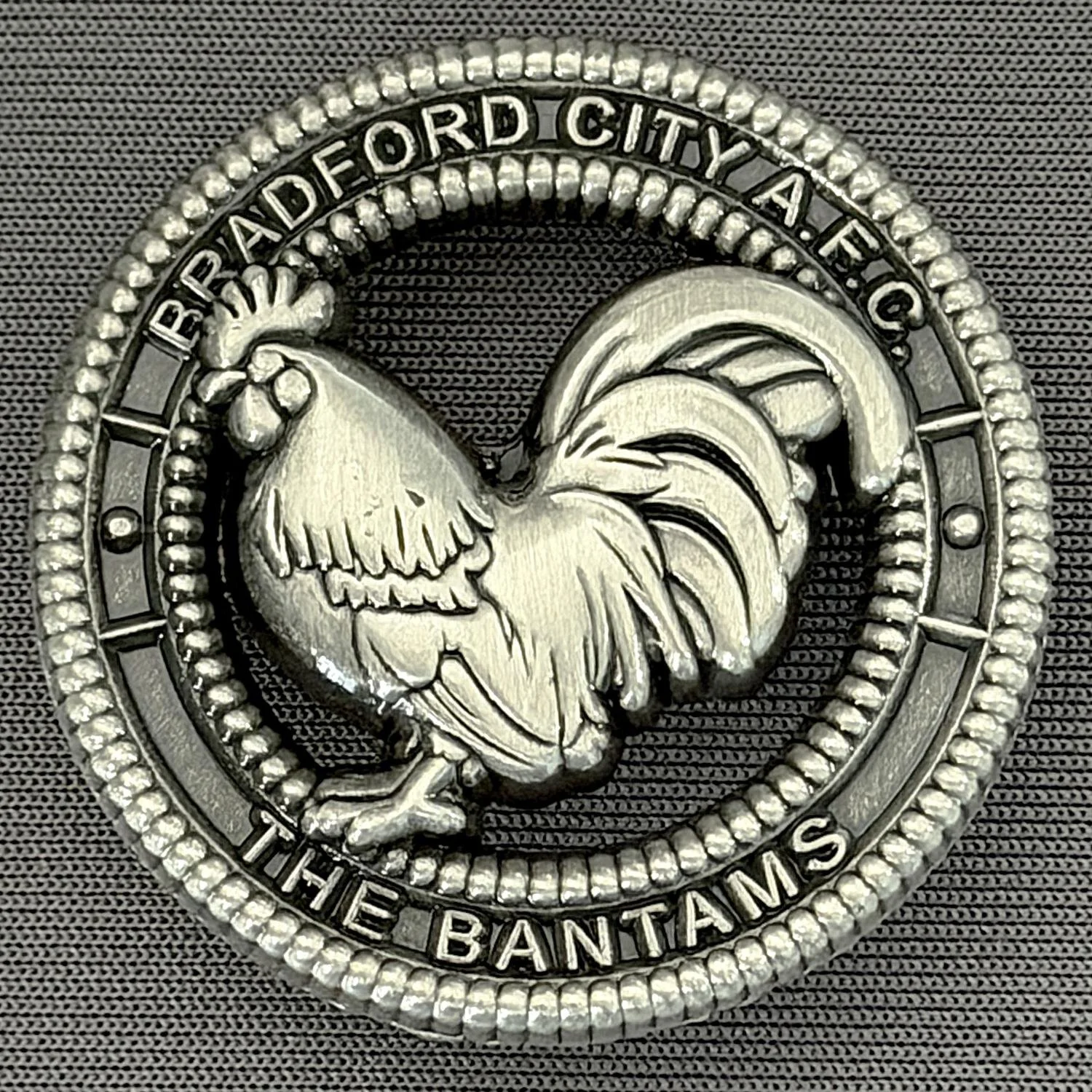 Close-up of a metallic badge or medal with a rooster in the center, surrounded by a beaded border. The words "BRADFORD CITY A.F.C." are at the top, and "THE BANTAMS" at the bottom.