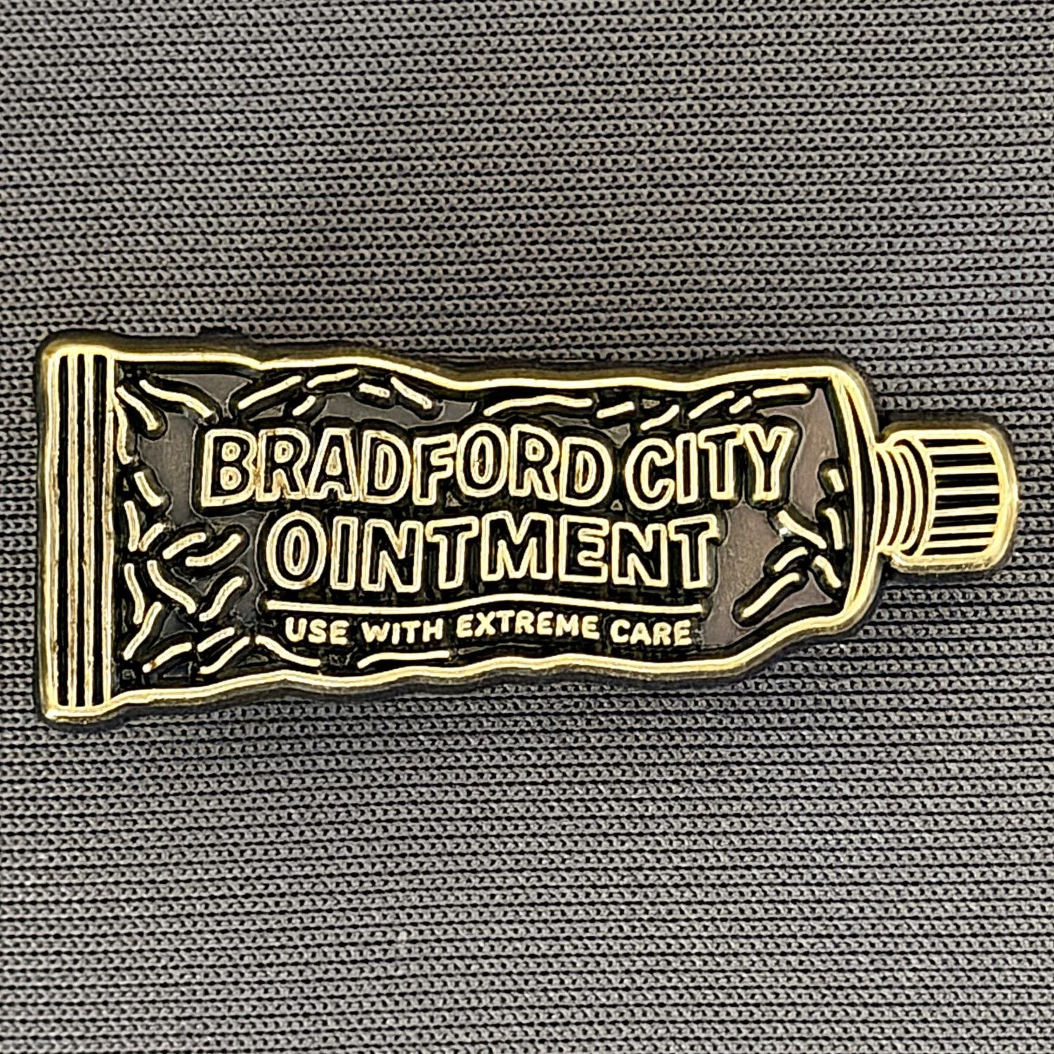A gold-colored badge shaped like a tube of ointment with the text 'Bradford City Ointment' and 'Use with Extreme Care' on a textured gray surface.