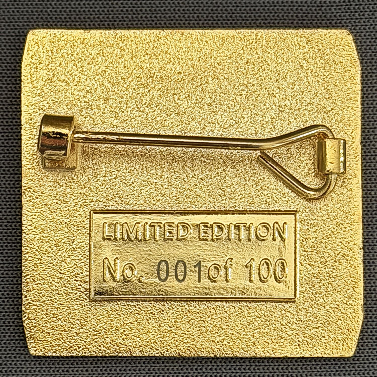 Gold-colored pin in the shape of a safety pin with a textured background. The pin has a rectangular label with the text 'LIMITED EDITION' and a serial number 'No. 001of 100' underneath.